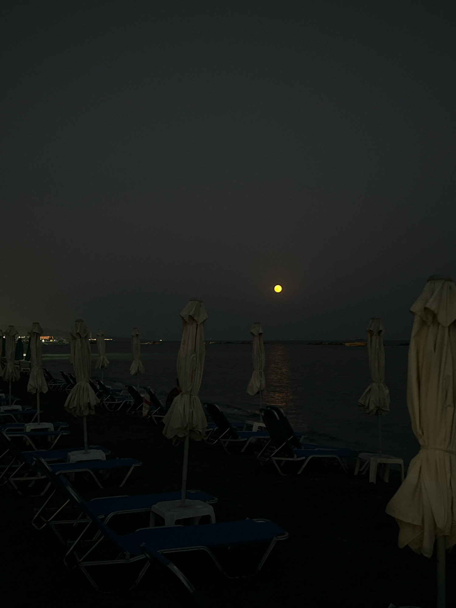 The image depicts a beach at night in Agios Tychona Community, Cyprus. A full moon is visible in the dark sky, casting a faint glow on the tranquil sea. Rows of closed beach umbrellas and empty lounge chairs line the sandy shore. In the distance, faint lights suggest buildings or other structures. The overall atmosphere is one of quiet serenity, with the moon and sea being the only prominent natural elements.