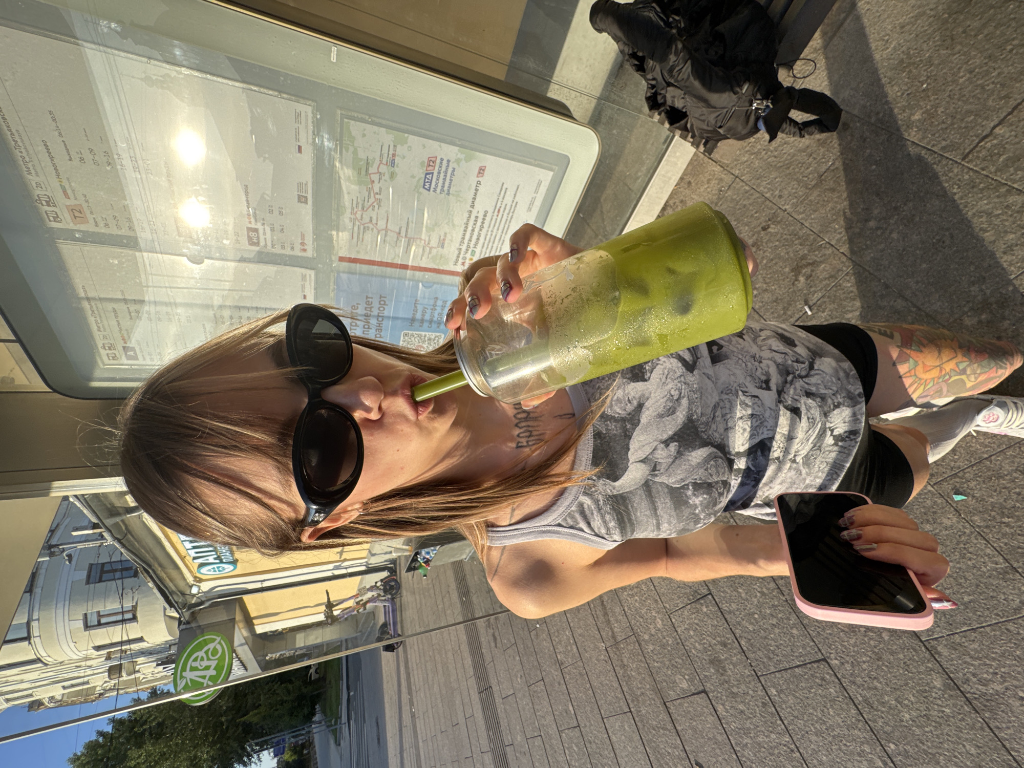 An overhead, medium shot captures a young woman standing outdoors, holding a pink smartphone in her left hand and a large, clear plastic cup filled with a green beverage and ice in her right. She is wearing sunglasses, a grey tank top with a monochrome graphic print, black shorts, and white socks. The woman is taking a sip from her drink through a straw. Her hair is light brown and falls past her shoulders.
The background reveals a sidewalk made of grey paving stones, a building with cream-colored walls and windows, and a glass display panel, likely at a bus or tram stop. The display panel contains information in Russian, including what appears to be a transportation map and schedule, with the MTA logo visible. In the distance, a purple electric scooter is parked. Sunlight casts shadows on the sidewalk, indicating it is daytime. The overall scene suggests an urban setting, likely in Moscow, Russia, given the Russian text on the display.