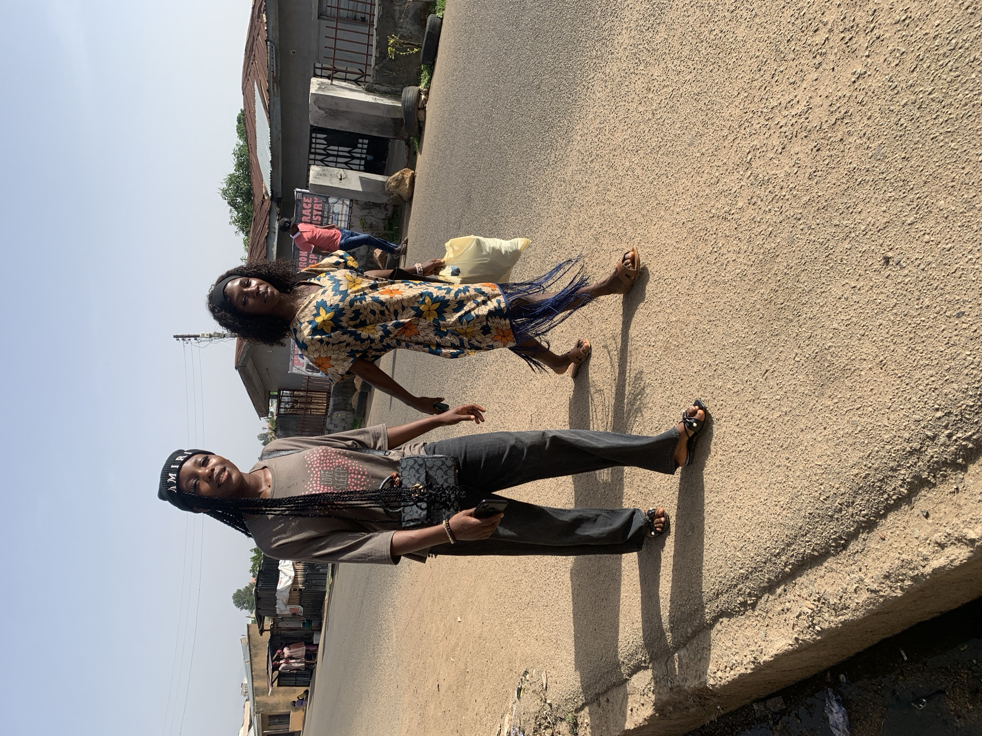 Two women stand on a paved road. One woman wears a black beanie, a grey shirt, dark pants, and sandals. She holds a mobile phone in her left hand, and a purse hangs over her shoulder. The second woman wears a floral print dress with fringe detail and sandals. She carries a yellow bag. The setting appears to be a street in Jos, Nigeria, based on architectural styles and environment visible in the background.