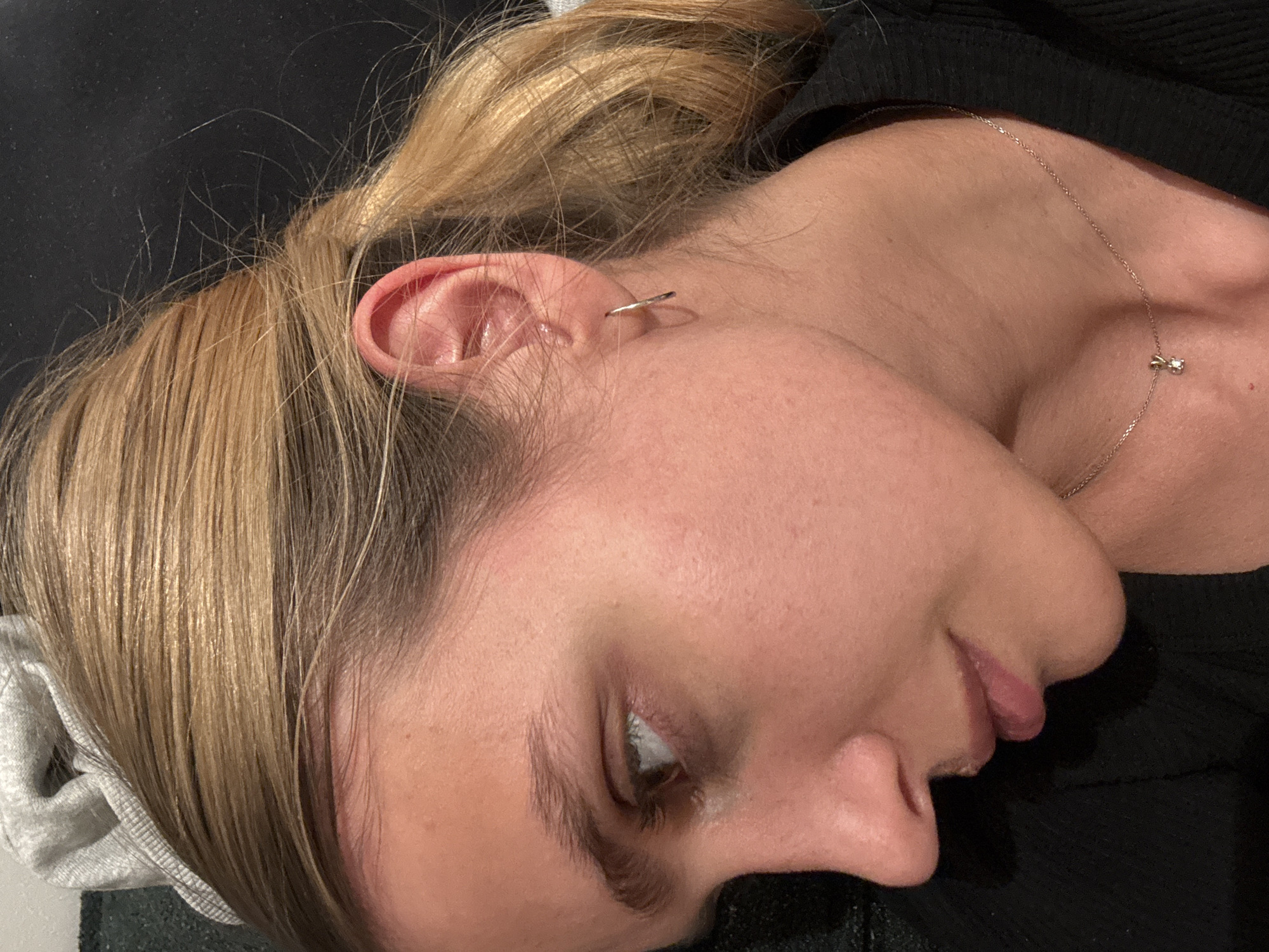 A close-up shot of a person's face, turned to the right, with the right ear and eye visible. The person has light blonde hair, pulled back into a ponytail. They are wearing an earring in their ear and a necklace with a small pendant. The background consists of a dark fabric. The location is unknown.