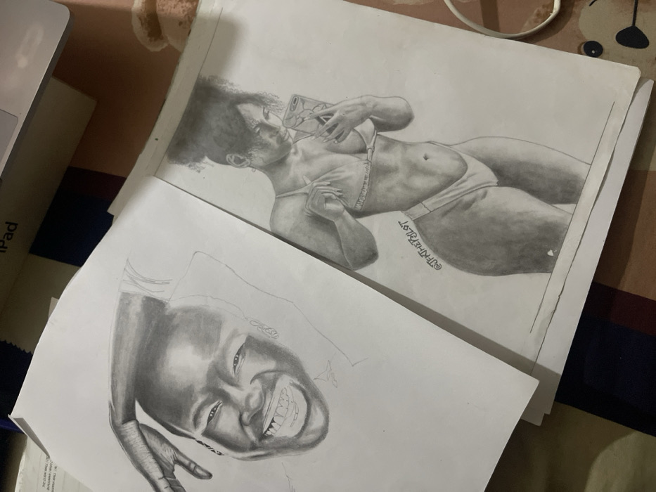 The scene, situated in Jos, Nigeria, displays two monochromatic pencil drawings on white paper, resting on a striped textile surface. The drawing on the left features a detailed portrait of an individual, likely male, depicted with a wide smile revealing teeth, and their left hand resting behind their head. The drawing on the right portrays a woman with an afro puff hairstyle, wearing a two-piece bikini, holding a smartphone in her left hand as if taking a mirror selfie, while her right hand adjusts her top. This drawing includes the visible watermark "@JFKTHEPYLOT". Partially visible in the upper background are a white electronic device with "iPad" visible on its casing, a dark colored laptop, a white power cord, and a light brown, plush object, all resting on the same striped textile surface.
