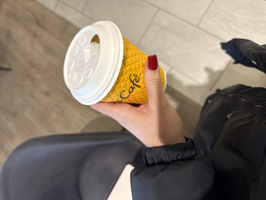 The image depicts a close-up, downward-angled view of a person's right hand holding a McCafé disposable coffee cup inside an establishment in Kyiv, Ukraine. The hand, with manicured nails painted dark red, grasps a yellow, textured paper cup prominently featuring the black "McCafé" logo. The cup is topped with a white plastic lid that has a cutout opening for drinking and various embossed circular and 'M' shapes.
The person is wearing a dark, likely black, puffer jacket, parts of which are visible in the lower left and bottom center of the frame. The background consists of interior flooring, transitioning from a lighter, possibly tiled surface on the right and bottom to a warmer, wood-grain patterned surface on the upper left. A dark grey, upholstered chair and a small portion of a white table are visible on the left side of the scene. The presence of the McCafé branding indicates the location is a McDonald's restaurant or a dedicated McCafé outlet within Kyiv. A specific address or neighborhood in Kyiv cannot be determined from the provided visual information alone.