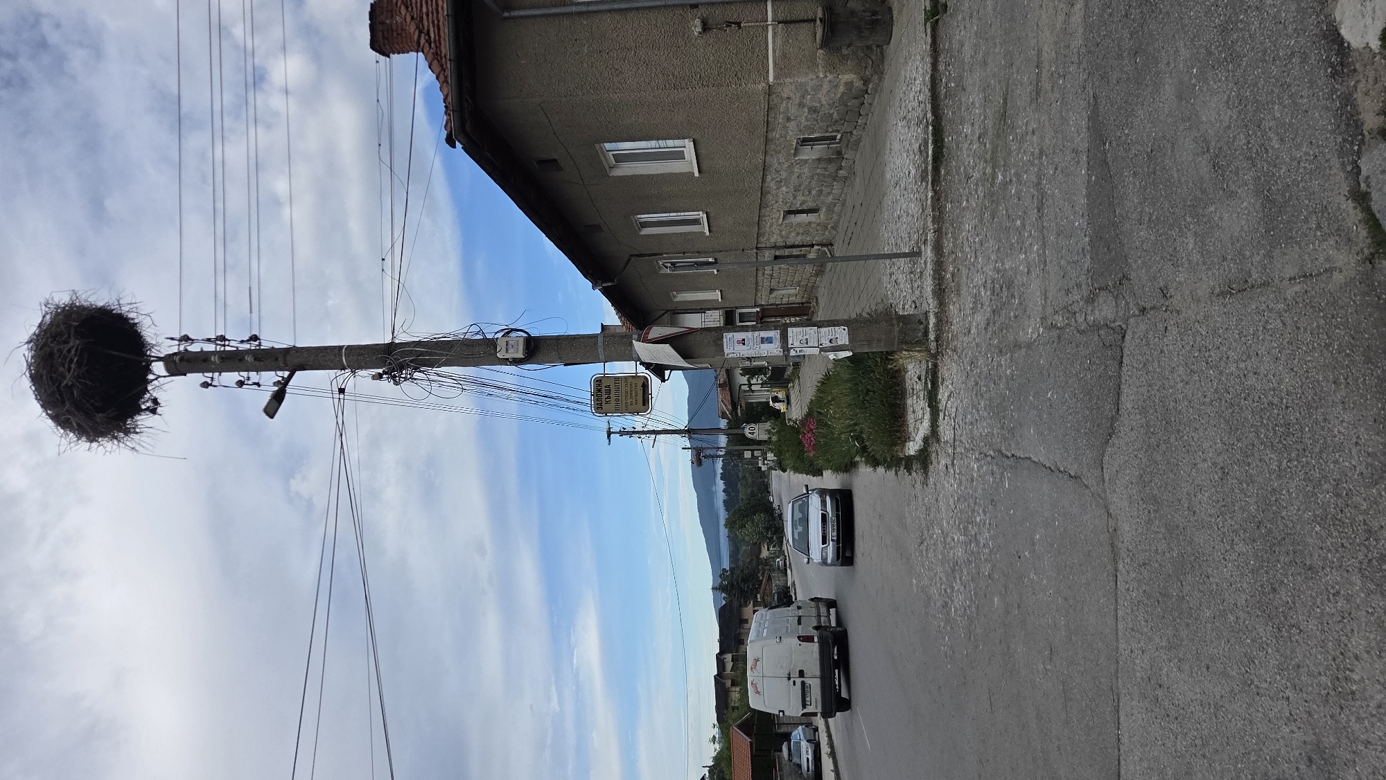 This image depicts a street scene in a village, likely in an Eastern European country given the Cyrillic signage. A utility pole with a large bird's nest at its apex dominates the foreground. The pole is adorned with numerous electrical wires, a street lamp, and several signs, one of which displays Cyrillic text and an arrow pointing to the right. To the right of the pole, a two-story building with a grey, textured facade and a red-tiled roof is visible, its ground floor constructed with stone. A sidewalk paved with irregular stones runs alongside the building.
The street itself is paved with asphalt, showing signs of wear and cracking. Two vehicles are present on the street: a white van on the left and a silver sedan further down the road. Several other vehicles are parked or driving in the distance. A speed limit sign indicating "40" is posted along the roadside. The background reveals more residential buildings, trees, and a body of water, possibly a lake or a bay, with mountains in the far distance. The sky is partly cloudy. The scene appears to be a quiet, everyday moment in a rural or semi-rural setting.