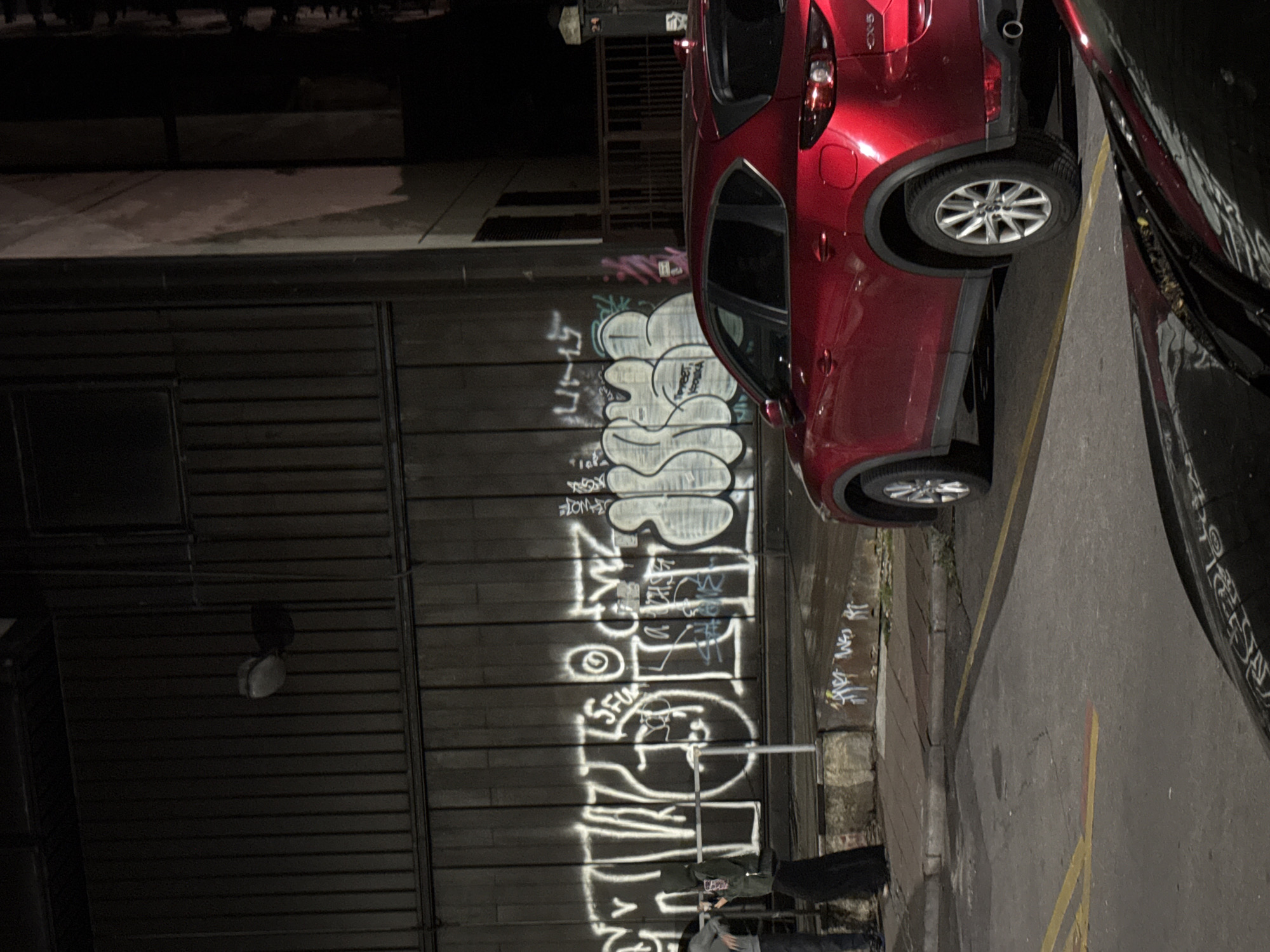 The image depicts a dark, urban street scene at night. The primary focus is a red Mazda CX-5 parked on the right side of the frame. The car is facing away from the viewer, with its rear end visible. The street is paved, with yellow and red painted lines indicating parking or traffic direction.
On the left side of the image, a dark metal wall is covered in white graffiti. Two individuals are standing near the wall. One person, wearing a dark hooded jacket and a long dark skirt, is looking down at their phone. Another person, partially visible, is wearing a grey hoodie.
The environment suggests a somewhat run-down or industrial area, given the corrugated metal wall and the presence of graffiti. A lamppost is visible on the wall, casting a shadow. In the background, a dark building or structure is partially visible, and a metal fence can be seen to the far right. The overall lighting is dim, typical of a nighttime urban setting. The graffiti on the wall appears to be stylized lettering, typical of street art. There is some minor debris or dirt on the ground and on the concrete barrier in front of the wall.
While the specific location in Belgrade, Serbia, cannot be definitively determined from the visual information alone, the presence of graffiti, urban architecture, and a parked vehicle are consistent with many city environments.