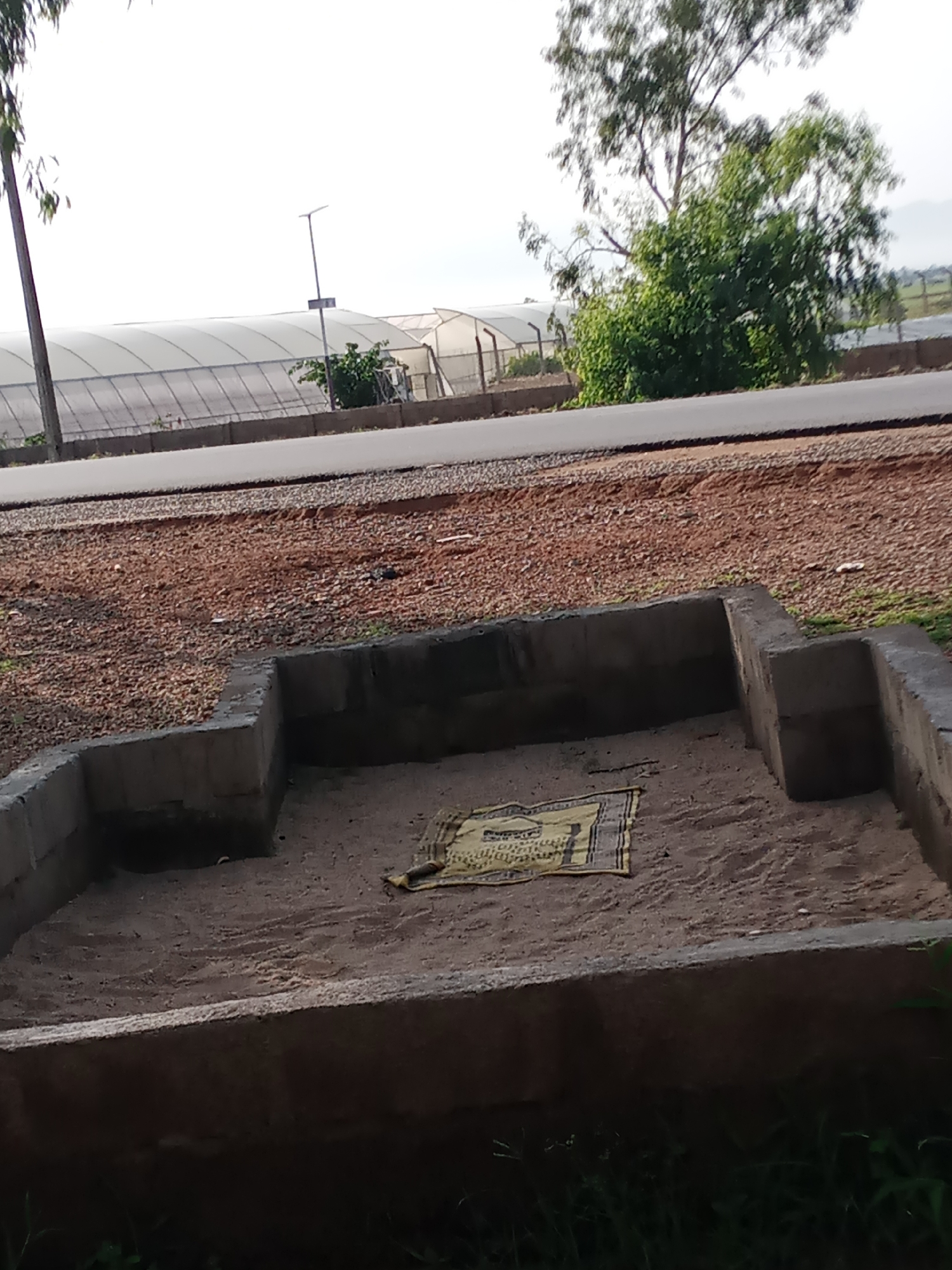 In Jauro Yinu, Nigeria, an outdoor scene features a small, rectangular, concrete-lined pit filled with sand. A yellow and black patterned mat is laid out in the sand, towards the left side of the pit. In the background, a paved road runs horizontally across the image. Beyond the road, large white greenhouses are visible, and further in the distance, a treeline and hills can be observed under a bright, overcast sky. A solitary street light stands near the greenhouses. The overall environment appears to be a semi-arid or rural area with agricultural structures.