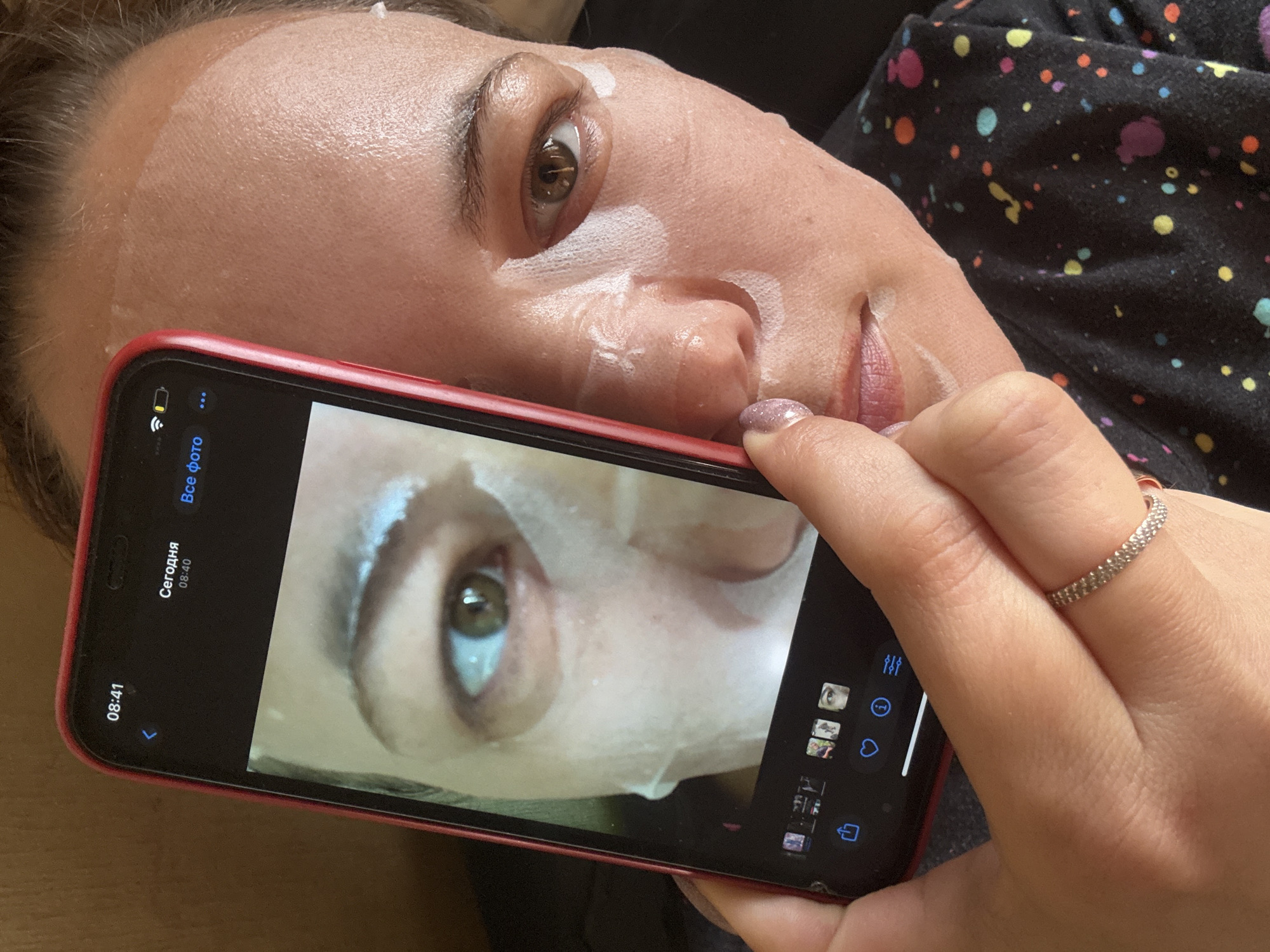 A woman is lying on her back wearing a sheet face mask, holding a red iPhone close to her face, displaying a photo of her eye and part of her face with the mask on. The iPhone screen shows the time 08:41 and the word "Сегодня" (Today) and 08:40 in Russian. The woman is wearing a sparkly ring on her hand. The backdrop is a dark fabric with colorful spots. There is no location in Dmitrov, Russia, visible in this photo.
