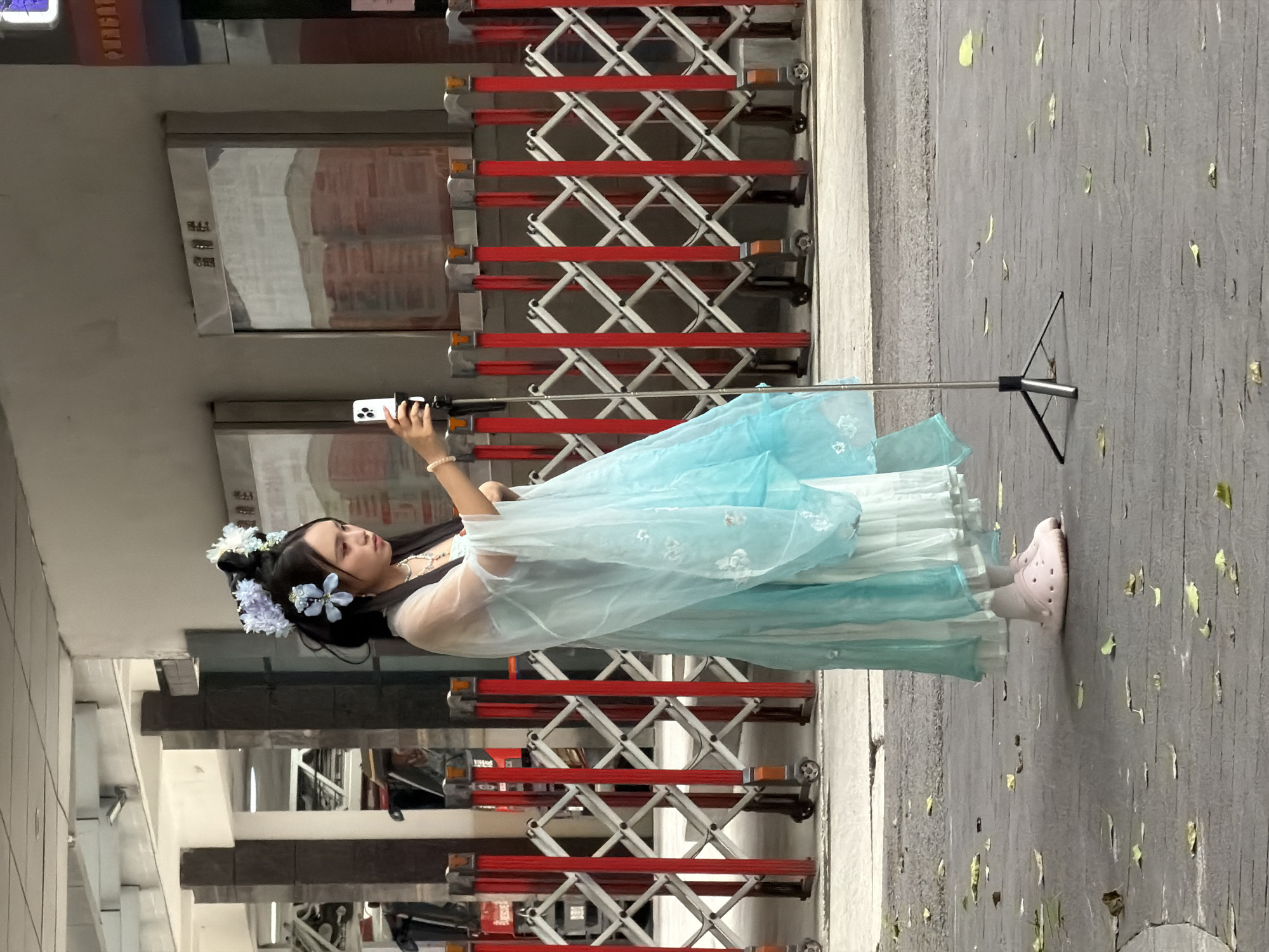 This media file, captured in Yuzhong District, China, features a young woman dressed in traditional Chinese Hanfu attire. She is wearing a light blue and white layered dress with delicate floral embroidery on the sheer outer layer. Her hair is styled with dark hair and adorned with white and blue flowers. The woman is holding a smartphone on a selfie stick, seemingly taking a photo or video of herself.
The scene appears to be outdoors, likely in an urban environment. Behind her are retractable metal gates, painted red and white, which are often used to block off areas. The ground is paved and scattered with fallen leaves, suggesting it might be autumn. The lighting indicates it is daytime, with natural light illuminating the scene. The woman's expression is neutral, focused on her activity. The overall atmosphere is that of a leisurely moment, perhaps a photo opportunity for the woman to capture her cultural attire. No visible text is discernible in the image.