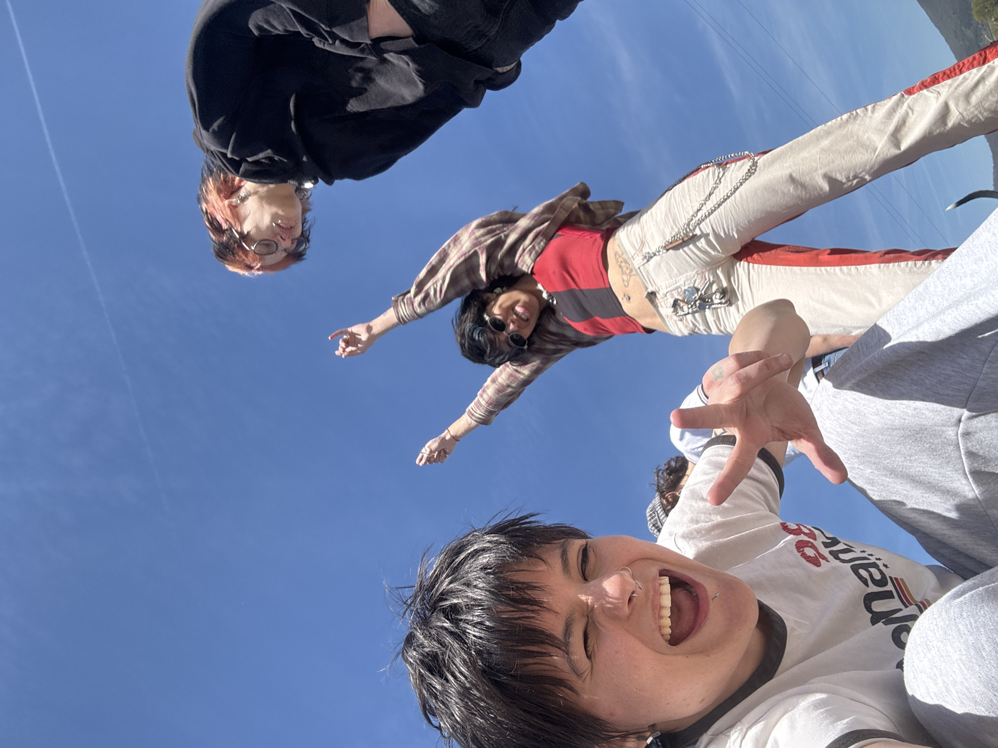 A low-angle shot of a group of five people against a clear blue sky, taken outdoors. At the bottom of the frame is a person with dark hair, wearing a white shirt with black trim, laughing and holding up one hand with fingers splayed. Above them, a person in gray sweatpants and a white shirt with black trim is partially visible. Further up is a person wearing cream-colored pants with red stripes, a red and black striped top, and a brown plaid jacket, with arms outstretched to the sides. Above and to the left, a person in a black hoodie with orange hair looks down. The location is not possible to determine based on this image, nor the city of Zhivkovo, Bulgaria.