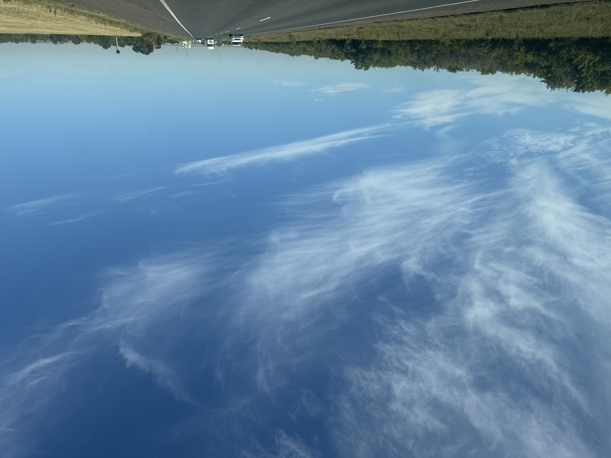 The image captures a highway scene under a vast, bright blue sky filled with wispy white cirrus clouds. The perspective is from a vehicle traveling on the road, looking forward. A dense line of green trees and vegetation borders the left side of the highway, while the right side features a harvested field of dry, golden grass. Several vehicles, including cars and trucks, are visible on the road ahead, indicating active traffic. The overall environment suggests an outdoor, daytime setting in a rural or semi-rural area. Based on the provided context, this scene is located in the city of Sorochinsk, Russia.