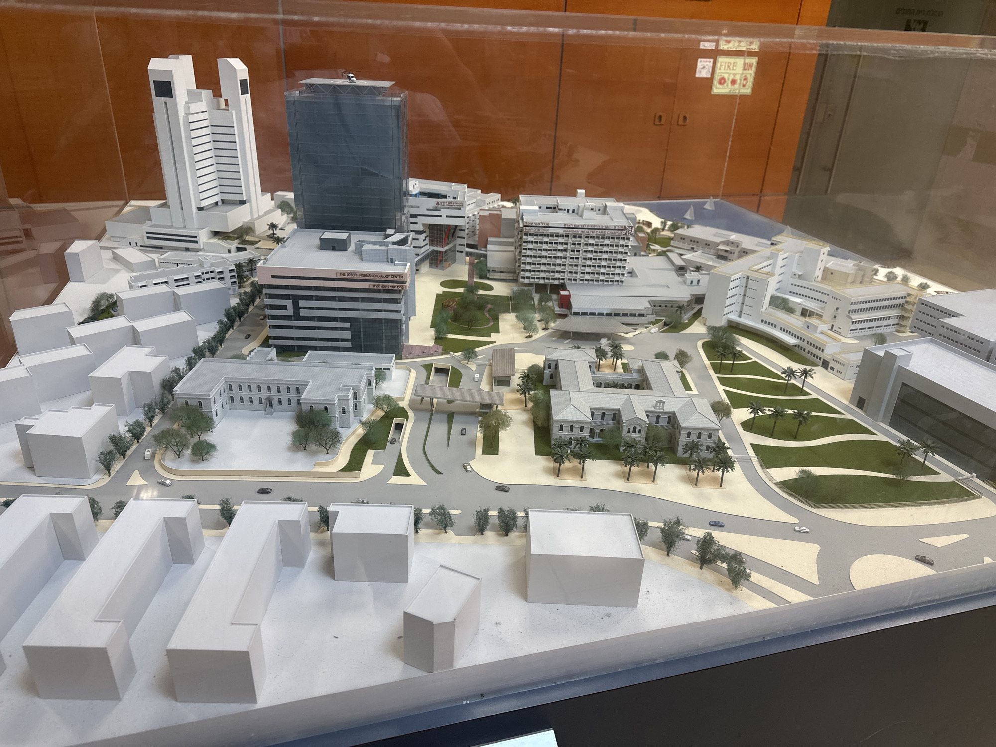 This is a detailed architectural model of a city complex, likely a hospital or university campus, situated in Haifa, Israel. The model showcases a variety of buildings, including modern high-rises with dark glass facades, and older, more traditional structures with arched windows and light-colored stone exteriors. Green spaces with trees and winding pathways are integrated throughout the model, suggesting a well-planned urban environment. Several miniature cars are placed on roads, and one building is labeled with Hebrew text and "The Joseph Fishman Oncology Center," indicating its function. A helipad is visible on top of one of the taller buildings. The model is enclosed within a clear display case, with reflections visible on its surface.