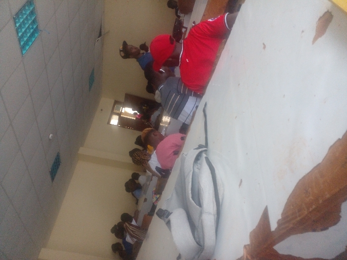 A classroom scene shows rows of students seated at desks. Most appear to be facing forward, likely towards an instructor. A person in a red shirt and cap is visible in the foreground. The room is bright, with light-colored walls and a ceiling with square panels. A doorway is seen in the background. The desks are white and show signs of wear. The image is rotated.