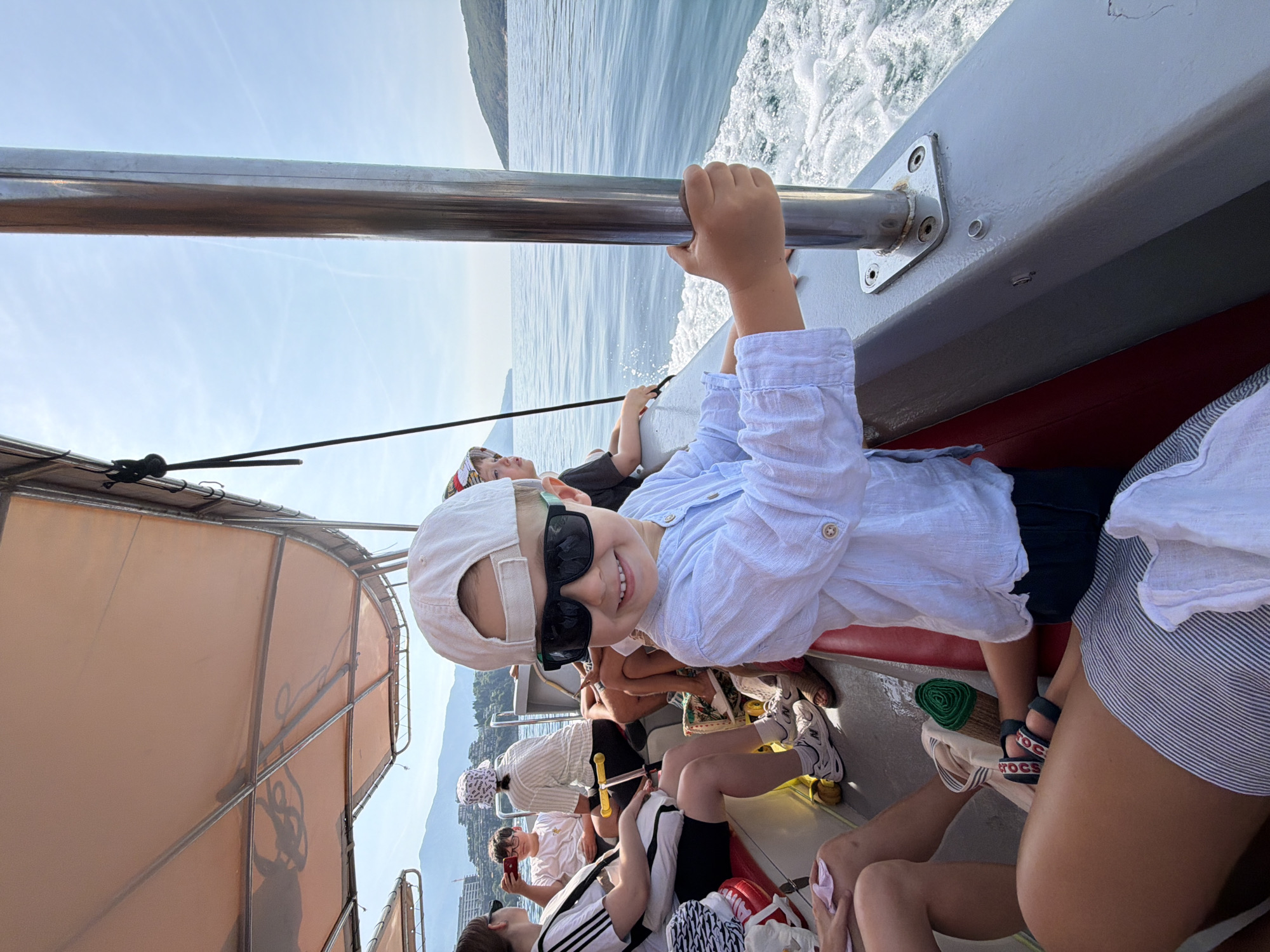 A medium shot captures a young boy in a white cap and sunglasses, wearing a light blue collared shirt and dark shorts, smiling as he holds onto a metal pole on a moving boat. Several other people, including adults and children, are seated behind him on the boat. The boat is on the water, with a mountainous coastline and buildings visible in the distance. The scene appears to be in the city of Budva, Montenegro.