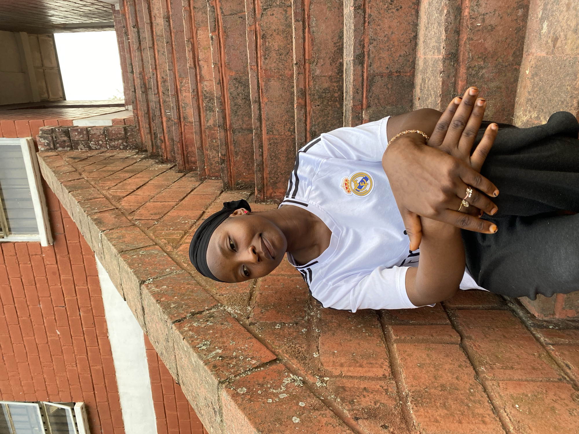 The image, taken in Vom, Nigeria, features a young person, likely female, with dark skin, positioned on a red brick surface. She is wearing a white long-sleeved shirt with black stripes on the sleeves and a black head covering. The shirt displays the crest of the Real Madrid football club on the left chest. She has a subtle, calm smile, and her hands are clasped in front of her. A gold-colored ring is visible on one finger, and a delicate gold-colored bracelet is on her wrist. Her fingernails have a reddish-brown color, possibly from henna.
The setting consists of weathered red bricks, forming what appears to be a staircase or a tiered platform. These bricks show signs of age, with some areas covered in moss or lichen. To the left in the background, a red brick wall with white-framed windows is visible. The upper background reveals a bright, open area, indicating an outdoor location under natural daylight, possibly diffused by an overcast sky or shade. The person is posing for the photograph in this casual, brick-built environment.