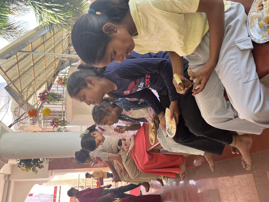 The image shows a group of five people, four women and one young girl, seated on a bench in an outdoor, covered area in Panchkula, India. The women are wearing colorful clothing, with one woman holding a smartphone and another eating something from a plate. The young girl is wearing a yellow shirt and is holding a piece of food in her hand. There is a plate with food in front of her. The people appear to be eating. They are seated on a brown bench with a white tile floor. The location is a covered outdoor area, possibly part of a temple or a community center, as indicated by the flowers and greenery in the background. The scene is lit by natural sunlight, suggesting it is daytime.