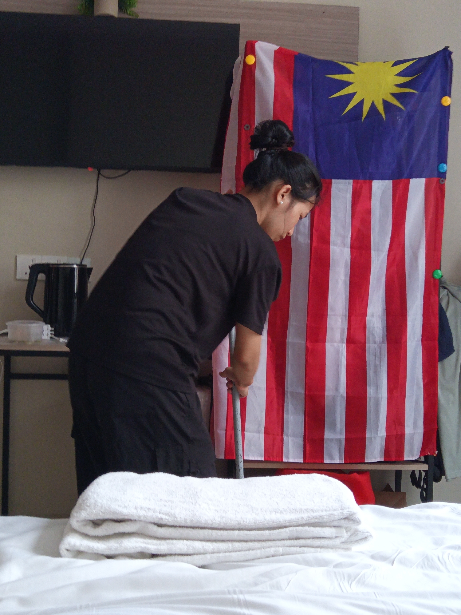 A woman in black attire is seen from behind, bending over to clean the floor with a broom. She has her hair tied up in a bun. Behind her, a Malaysian flag is displayed prominently. In the foreground, a stack of neatly folded white towels rests on a white surface, likely a bed. To the left, a black electric kettle is visible on a shelf, with a wall socket above it. A large black television screen is mounted on the wall in the upper left corner of the image. The setting appears to be an indoor space, possibly a hotel room or a residence in Kuala Lumpur, Malaysia, given the presence of the Malaysian flag.