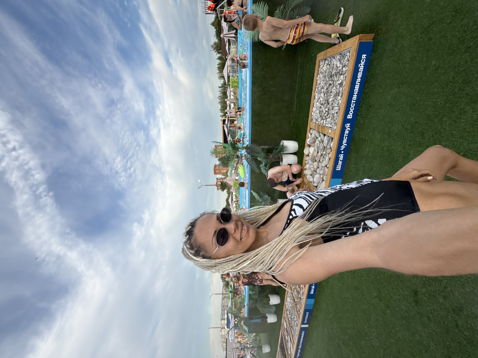 In Rusanovo, Russia, a woman with blonde braided hair and sunglasses is taking a selfie in front of a crowded outdoor swimming pool on a cloudy day. She is wearing a black and white patterned swimsuit. The pool area has several people swimming and relaxing under umbrellas. In the foreground, there are textured pathways with stones, labeled in Russian, indicating a wellness or sensory experience area. A young boy in striped swim trunks stands to the right, facing away from the camera, near one of the pathways. The overall scene depicts a recreational water park environment.