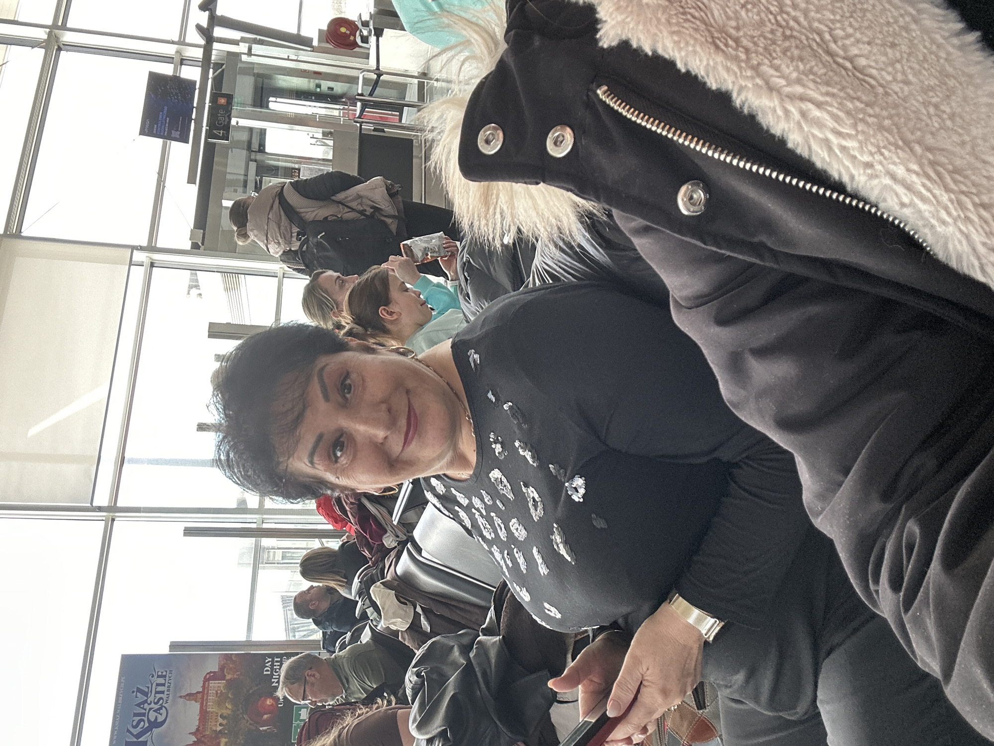 The image appears to be taken at Wrocław Airport in Wrocław, Poland. A woman with short dark hair is looking at the camera; she is wearing a black top with white patterned designs and a gold bracelet. The background shows a waiting area, with other people sitting and standing near large windows and gate signage ("Gate 4"). There is also promotional material visible, possibly an advertisement for Książ Castle. Some individuals are using electronic devices. The image is taken at an angle, possibly as a selfie.