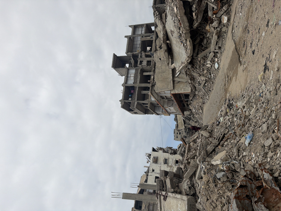 Rubble and collapsed buildings dominate the scene under a cloudy sky. Concrete debris is scattered across the foreground and background, indicating widespread destruction. The skeletal remains of structures rise from the debris, with exposed rooms and floors visible. The destruction suggests a war zone or a disaster area, conveying a sense of devastation.