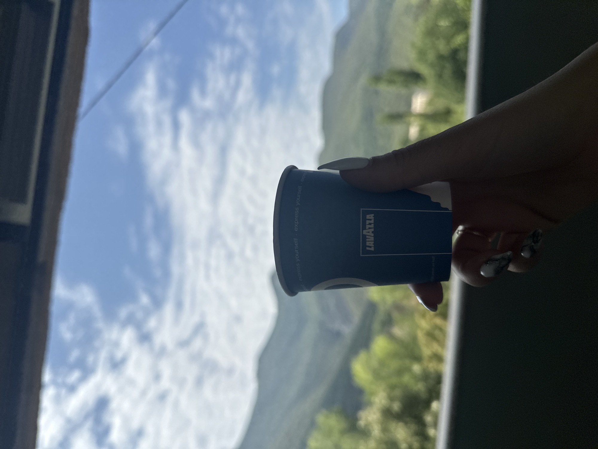 A hand holds a small blue paper coffee cup with the Lavazza logo. The cup is branded with white text that reads "express yourself". The background is a blurred landscape of green mountains under a bright blue sky with white clouds. The scene appears to be outdoors, possibly on a balcony or terrace, with a dark railing in the foreground. No specific location in Vratsa, Bulgaria, is discernible from the image.