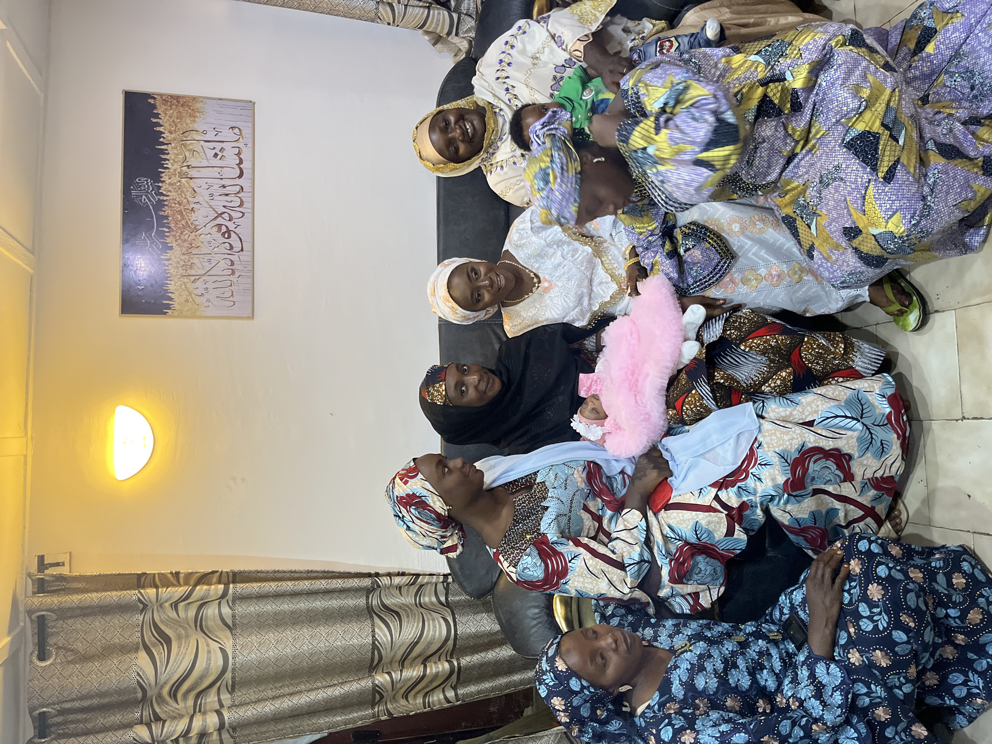 A group of seven individuals, five women and two babies, are gathered indoors, likely in a residential setting in Jos, Nigeria. The women are dressed in colorful traditional attire, featuring headscarves and patterned fabrics.
In the center of the image, a young baby in a pink garment is being held by a woman. Another baby, wearing a green shirt, is visible on the lap of a woman to the right. The women are seated on a black sofa.
The room appears to be a living area, with curtains visible on the left and a framed picture of Arabic calligraphy on the wall to the right. A wall-mounted light fixture illuminates the space with a warm yellow glow. The flooring is tiled. The overall atmosphere suggests a family gathering or a celebration.