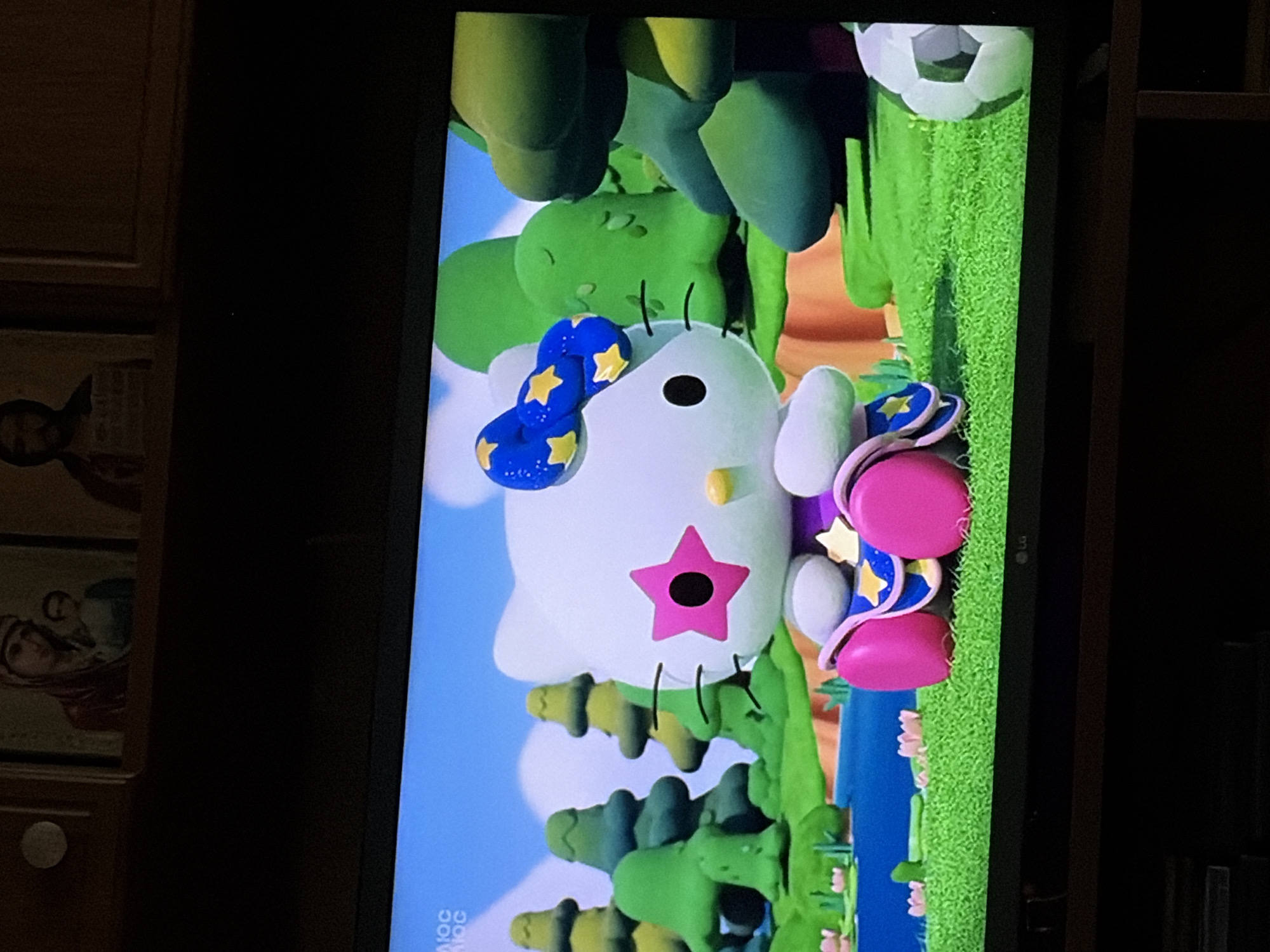 This is a medium shot of a television screen displaying a 3D animated scene of Hello Kitty in a whimsical, colorful outdoor environment. Hello Kitty, a white anthropomorphic cat with a pink bow on her head adorned with yellow stars, is sitting on a green, grassy surface. She is wearing a purple and pink outfit with a blue collar and yellow stars. Her eyes are black dots, and her nose is a small yellow oval. A large pink star with a black center is on her left cheek.
The background features a bright blue sky with fluffy white clouds. In the middle ground, there are several cartoonish, brightly colored green trees of varying sizes and shapes, some resembling oversized mushrooms or stylized evergreens. To the left of Hello Kitty, a calm blue body of water with some pink lily pads is visible.
In the far right of the screen, a black and white soccer ball is partially visible. At the top of the television screen, partially visible, are two framed religious icons. The left icon appears to depict the Virgin Mary holding the infant Jesus. The right icon is darker and less discernible, but seems to show a bearded figure. The overall scene is cheerful and playful. The television is placed on a dark wooden furniture piece. The setting is unknown.