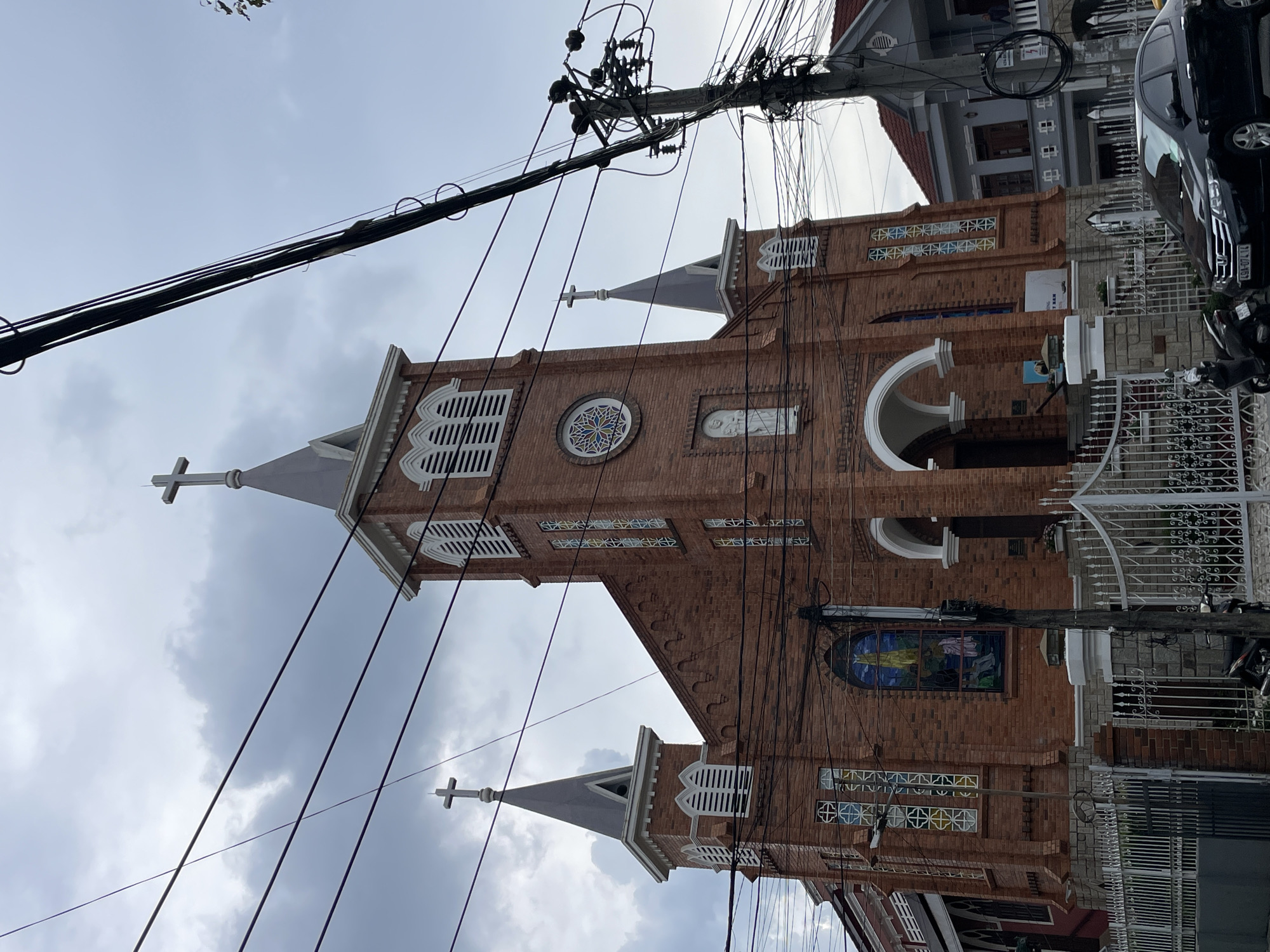 A brick church with two bell towers and pointed spires, each topped with a cross, dominates the frame. Stained-glass windows adorn the facade, and a statue of a religious figure is visible on the front tower. The church is partially obscured by a tangle of power lines and a utility pole in the foreground, which also features a street light. A white metal fence runs along the front of the property, with a gate. Beyond the church, a gray building with a red tiled roof is visible. A black sedan and a motorcycle are parked on the street in front of the fence. The sky is overcast and gray, suggesting cloudy weather. The church is likely the Domaine de Marie (also known as the Convent of the Sisters of the Lovers of the Holy Cross) in Da Lat, a city in the Lâm Viên plateau of Vietnam, known for its French colonial architecture and as a popular tourist destination.