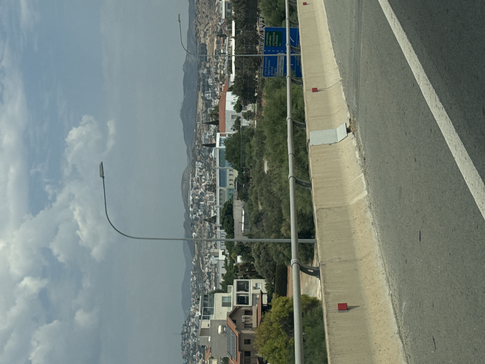The image depicts a view from a road, likely an overpass or highway, looking towards a densely populated area with buildings on hilly terrain. The sky is partly cloudy. In the foreground, a concrete barrier separates the road from the embankment. Red markers are visible on the barrier. Beyond the barrier, there are trees and some residential buildings, including one with solar panels. Further in the background, a sprawling urban landscape with numerous houses and structures climbs the hills. A street light is visible on the right side of the frame. A blue sign with text in Greek and English is also present on the right, indicating directions to various locations including "AGIA FANEROMENI", "LIMASSOL", "A1", and "TOWN CENTRE". The presence of the sign with Greek and English place names and the "A1" highway designation strongly suggests the location is within Cyprus, likely on a main road connecting to or within the Limassol district. Specifically, the place names on the sign, such as "Agia Faneromeni" and "Limassol", along with the typical Cypriot landscape of hills and dense housing, point towards the area of Kato Polemidia Municipality, which is part of the Limassol urban area.