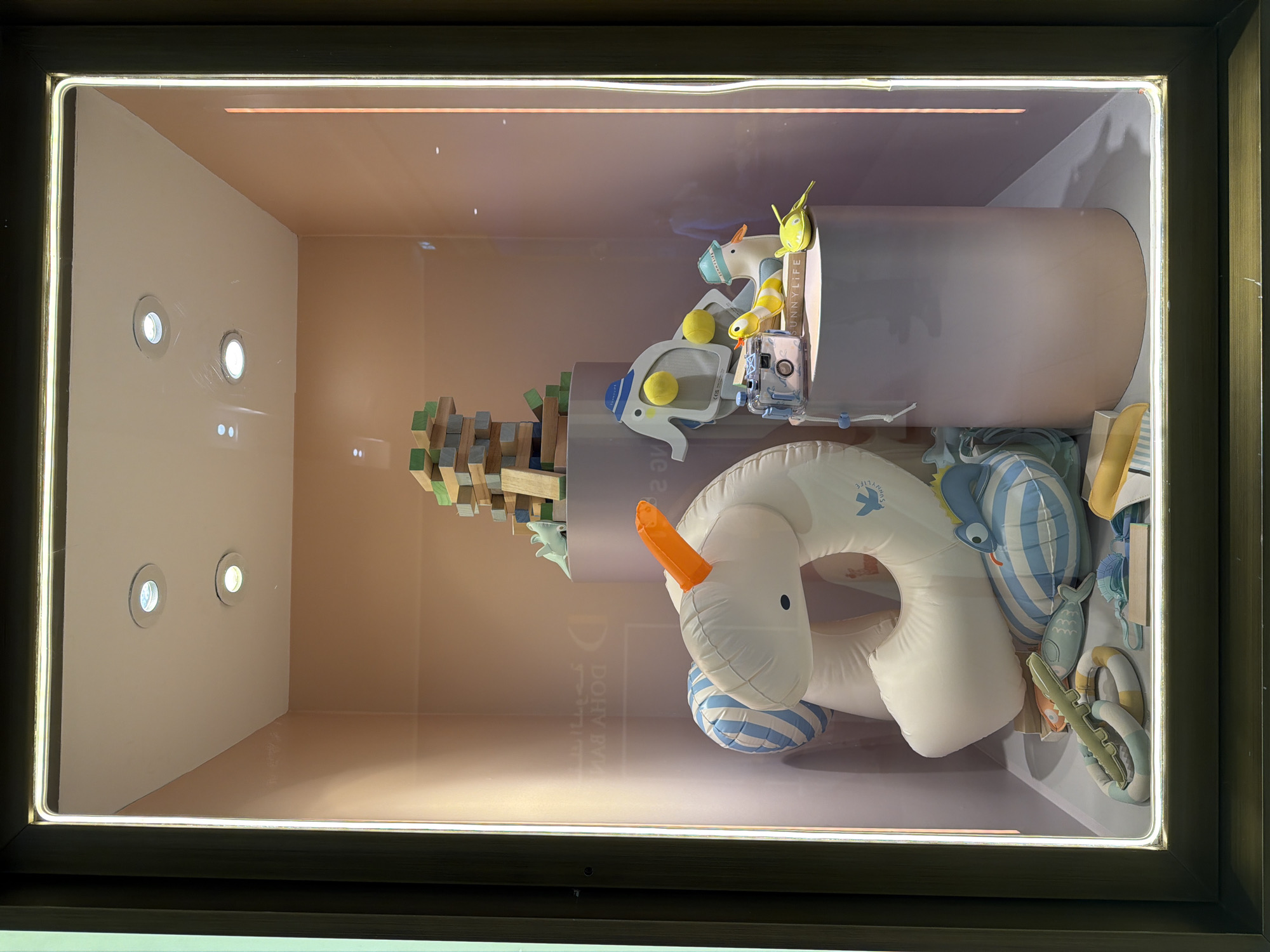 This is a medium shot, eye-level interior photograph of a retail display window, likely for children's toys. The display is set against a pale peach-colored wall and features several objects. In the center, a large, inflatable white unicorn with a bright orange horn is the focal point. To the left and slightly behind the unicorn, a blue and white striped inflatable shark is visible. In front of the unicorn and shark, on the floor of the display, are several small, colorful toys, including a green alligator-shaped object and two ring-shaped toys, one with blue and yellow stripes and the other with white and yellow stripes.
On the right side of the display, two cylindrical pedestals are present. The larger pedestal, in the background, holds a stack of wooden blocks, some of which are green. On the smaller pedestal in the foreground, a toy elephant with yellow balls for eyes and a blue hat sits. Next to the elephant is a toy duck with a blue hat and an orange beak, and a toy yellow and white striped snake. A small, transparent blue camera with a black strap is also placed on this pedestal, along with a green toy fish and a yellow toy boat. The entire display is enclosed by a glass window with a thin, illuminated white border around the perimeter. The background wall is slightly illuminated by the lighting within the display. The overall impression is a playful and whimsical arrangement of bath or pool toys. The location is identified as Unknown, Unknown.