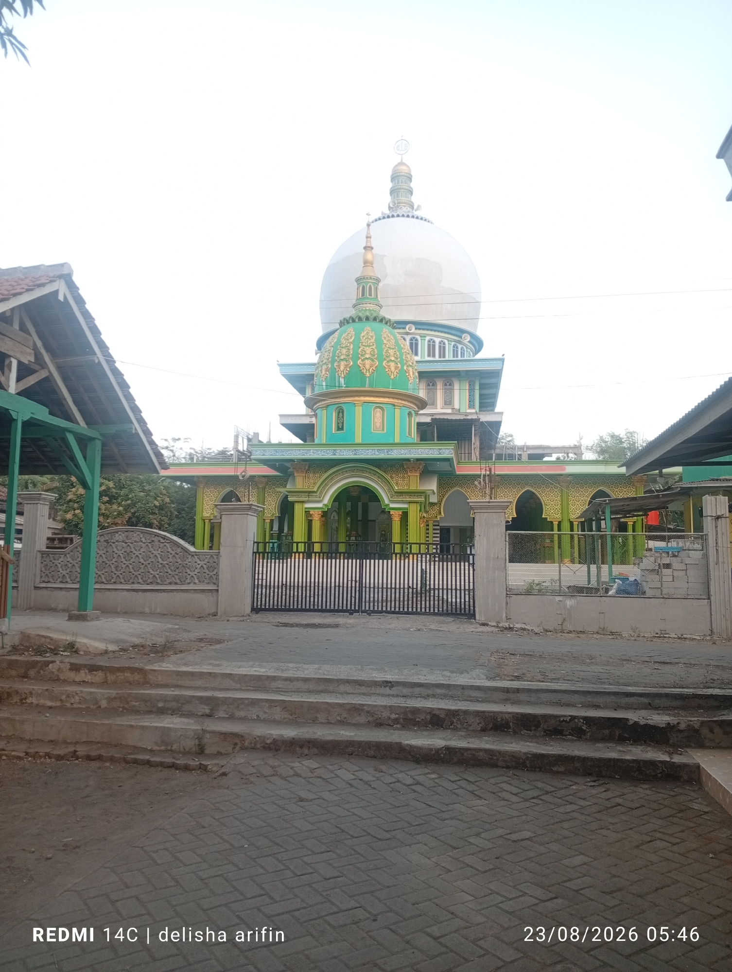 The image shows a mosque with a large white dome and a smaller green dome. The mosque has green and gold accents and is surrounded by a fence and wall. In the foreground, there are steps leading up to the mosque and a paved area. The sky is overcast. The text at the bottom of the image indicates the photo was taken on August 23, 2026, at 5:46 AM. The location is identified as Tamansari, Indonesia.