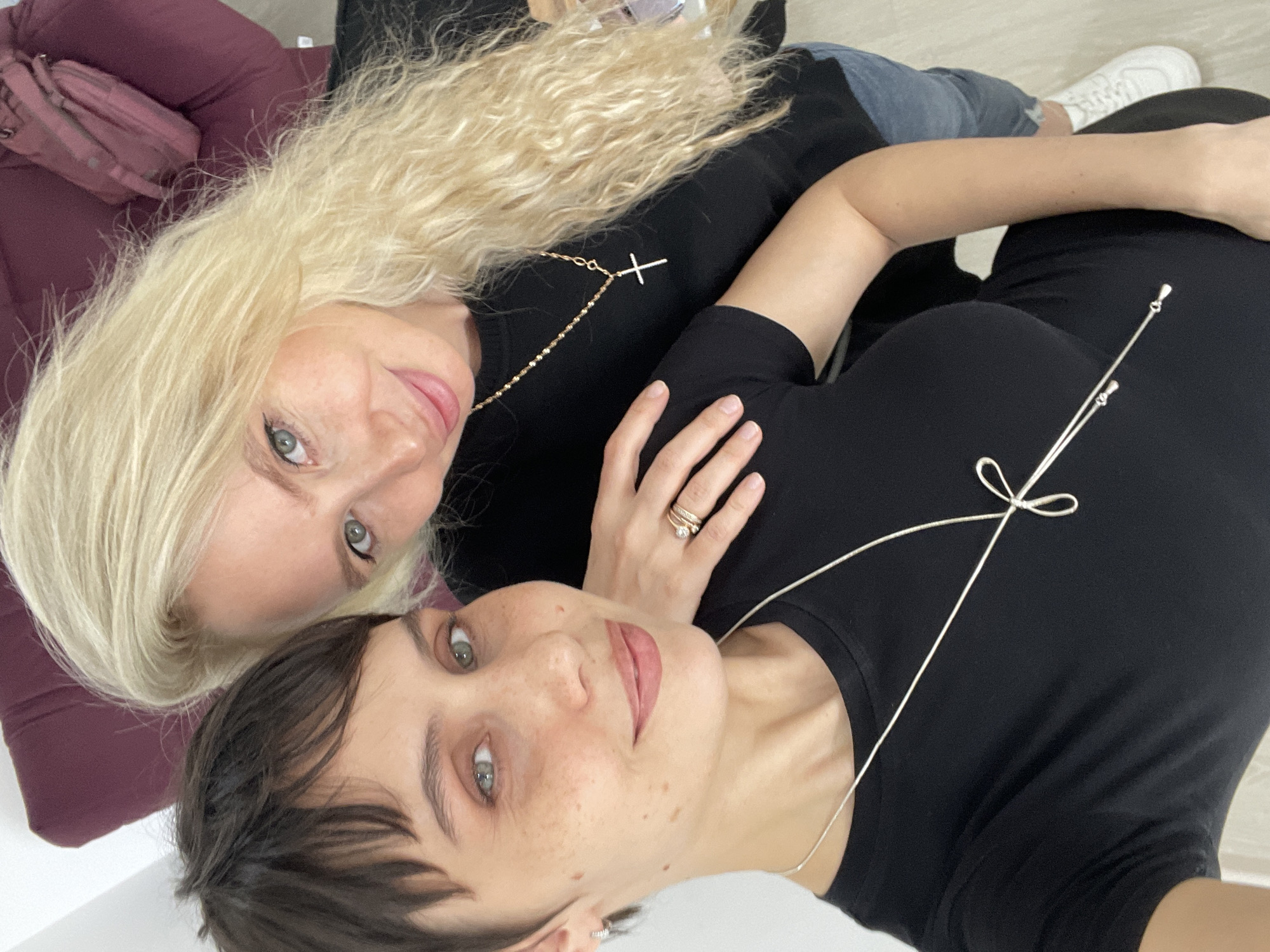 Two women are posing closely together for a selfie indoors. One woman has long blonde wavy hair and is wearing a black top with a gold cross necklace. The other woman has short dark hair, freckles, and is wearing a black top with a silver necklace. She also has rings on her hand. In the background, part of a maroon-colored cushion or bag is visible. It is not possible to confirm the specific location within the city of Izhevsk, Russia, based on the given image.