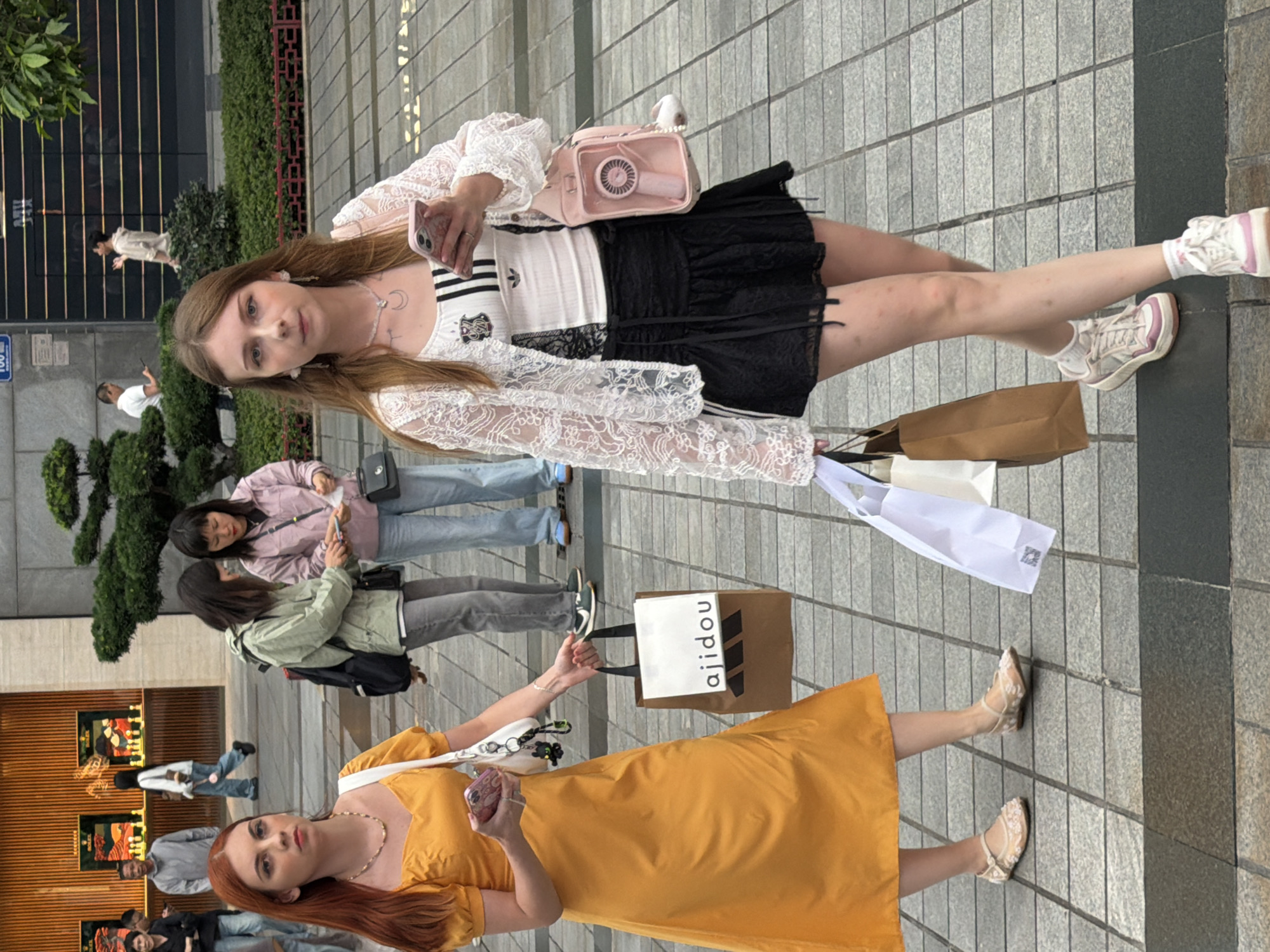 Two young women are walking on a paved sidewalk in Yuzhong District, China. The woman on the left has long red hair and is wearing a yellow dress and light-colored flats. She is carrying a white purse and a shopping bag with the logo "ajidou". The woman on the right has long light brown hair and is wearing a white lace top, a black skirt, and sneakers. She is holding a pink phone and a small pink bag with a fan attached. She is also carrying a shopping bag. In the background, there are other pedestrians and a building with storefronts. The environment appears to be an urban shopping area.