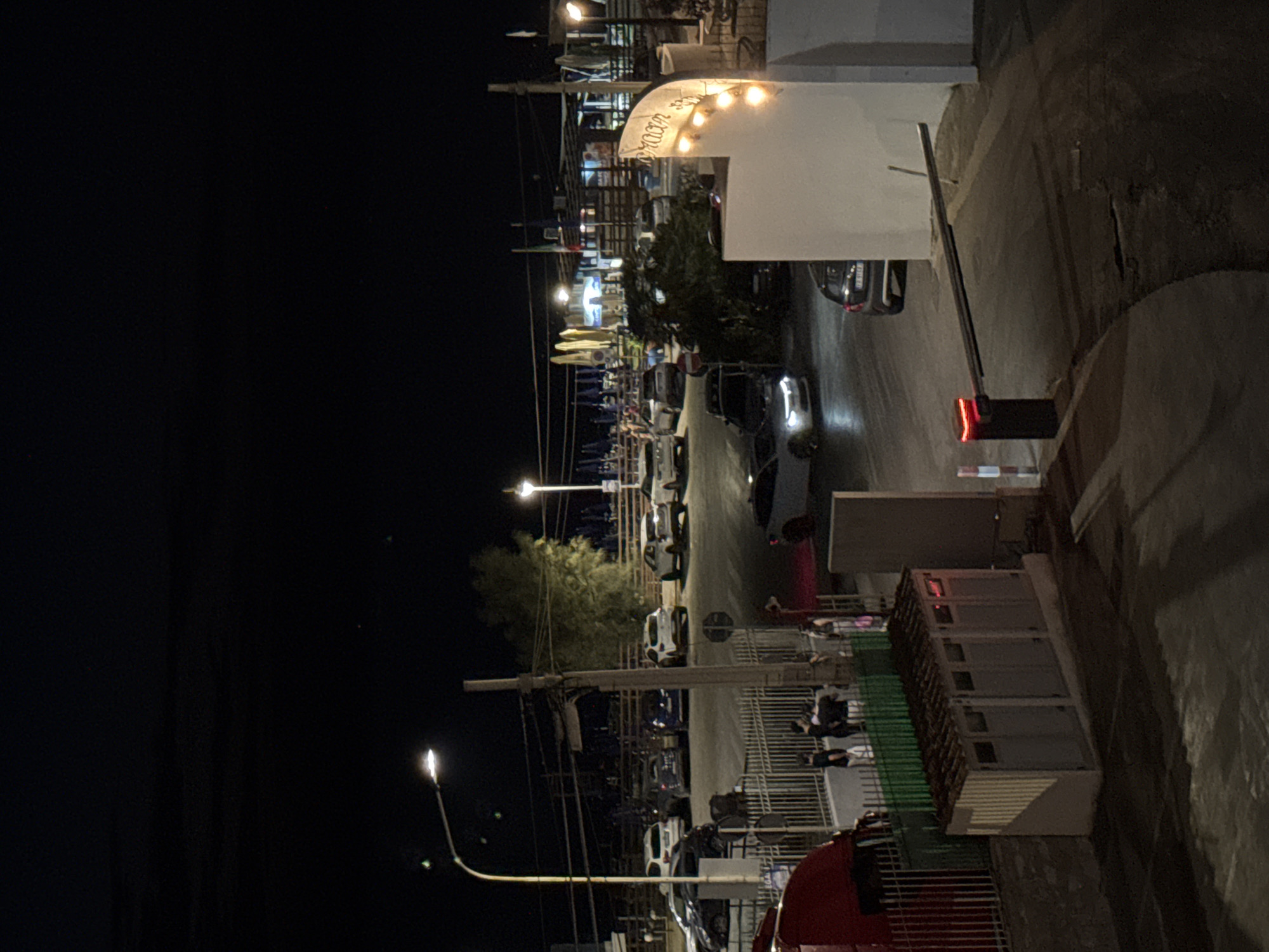 The image depicts a nighttime scene at what appears to be an entrance or exit to a facility, possibly a beach club or resort, in Capaci, Italy. In the foreground, a red vehicle, likely a truck or van, is partially visible on the left. Adjacent to it is a metal fence and a low building with multiple compartments, possibly utility structures or restrooms, featuring a corrugated roof. To the right of this building, a barrier gate with a red light is lowered, indicating a closed or controlled entry point. A white structure, possibly part of a building or wall, rises on the right side of the frame, featuring decorative string lights.
In the middle ground, several cars are parked or driving through the area. A white convertible is prominent in the center, and other vehicles, including SUVs and sedans, are visible. A group of people, dressed in casual attire, are gathered near the fence, some appearing to be talking or socializing.
The background reveals more of the facility, with what looks like beach umbrellas, tables, and more buildings illuminated by artificial lights. Several streetlights and building lights cast a glow on the scene, creating a contrast with the dark night sky. The overall impression is a lively but orderly environment at night. The presence of Italian flags in the background suggests a possible location within Italy, and the context provided specifies Capaci, Italy.