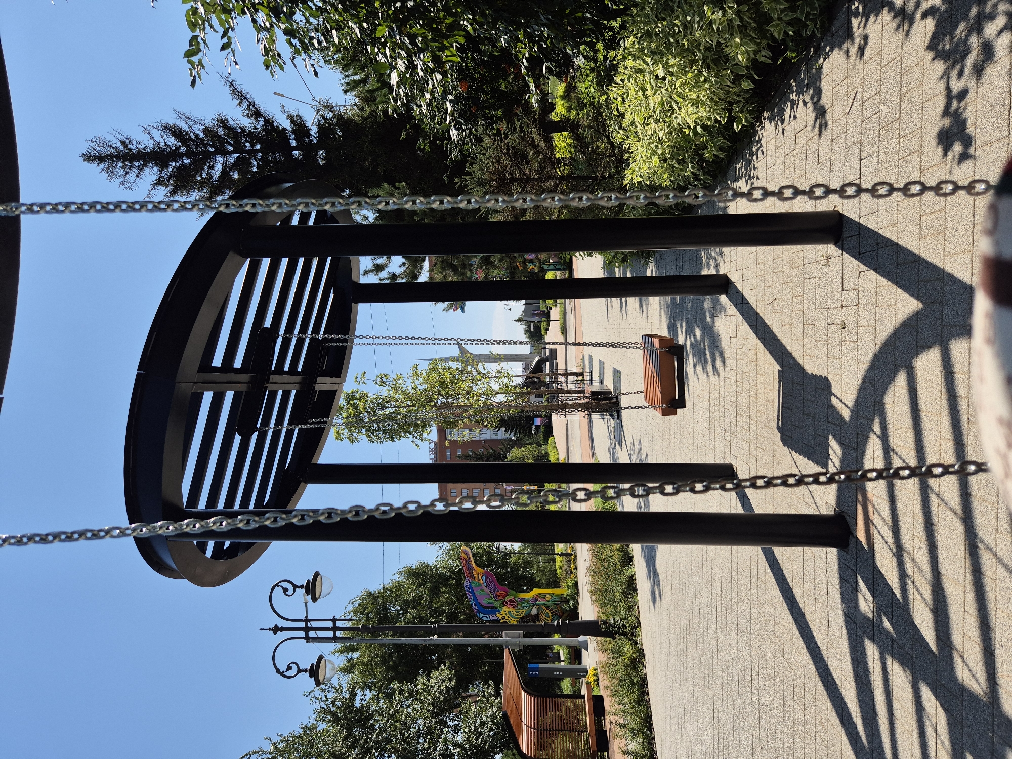 The image depicts a modern swing set structure with black metal framing and a circular roof. A wooden swing seat hangs from metal chains. The swing set is situated on a paved walkway made of light-colored rectangular stones. Tall green trees and lush bushes flank the walkway. In the background, a stylized sculpture of a bird with vibrant colors is visible, along with a street lamp and a covered bench. A multi-story building is also present in the distance. The scene is set under a clear blue sky on a sunny day, casting distinct shadows from the swing set onto the pavement. The location is the city of Kokshetau, Kazakhstan. There are no people present in the image.