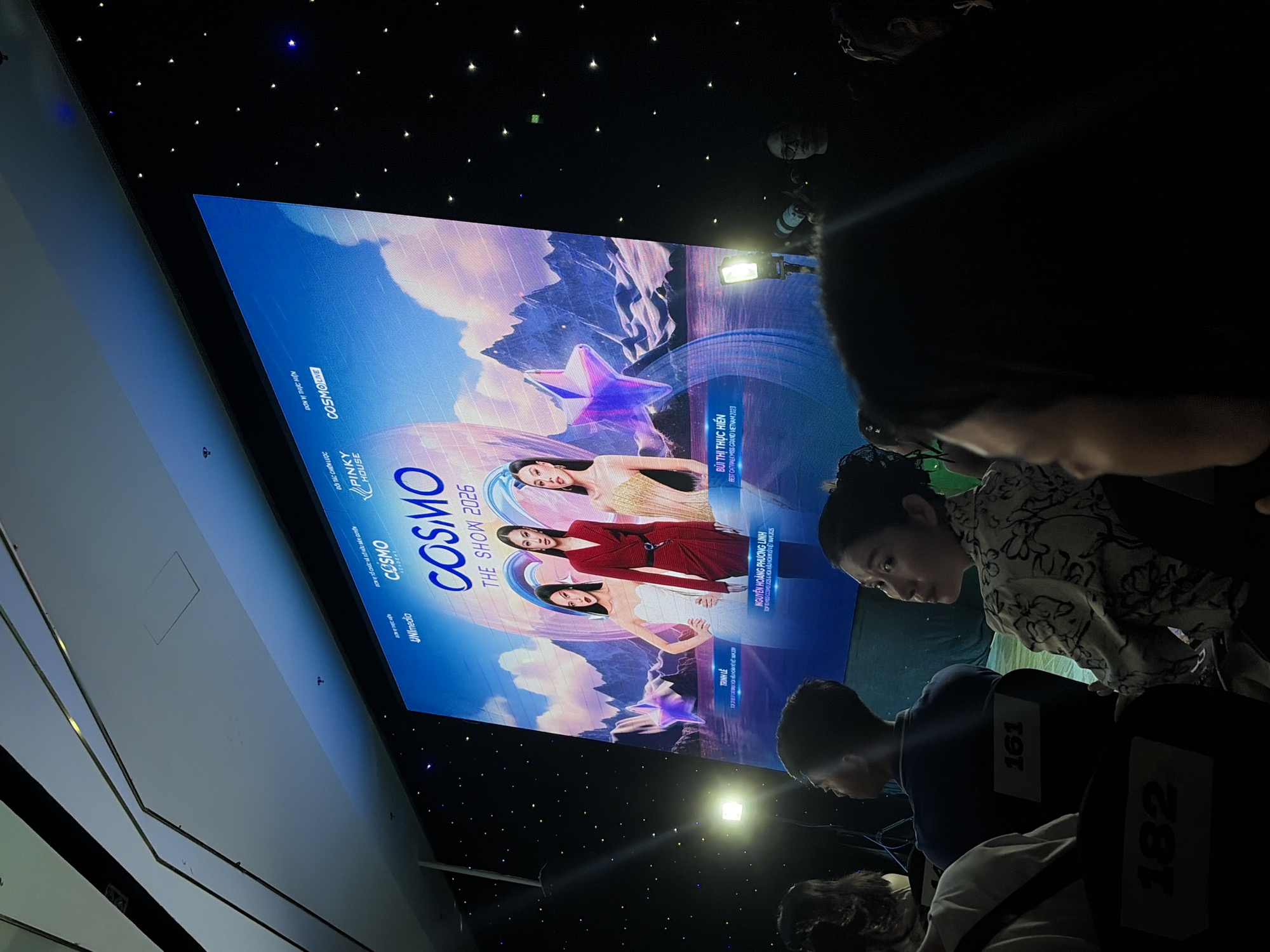 The image depicts an indoor event taking place within Nha Trang Ward, Vietnam. A large, brightly illuminated screen serves as the central focal point, displaying the title "COSMO THE SHOW 2026." Text identifying sponsors and partners, including "UNImedia," "COSMO ACADEMY," "PINKY HOUSE," and "COSMO LIVE," is visible on the screen. The main graphic features three female figures: "TRỊNH LÊ" on the left in a white gown, "NGUYỄN HOÀNG PHƯƠNG LINH" in the center in a red suit-dress, and "BÙI THỊ HIỀN" on the right in a gold strapless dress. The background of the screen graphic includes stylized mountains, a body of water, and prominent star shapes in shades of blue and purple.
The event space is dimly lit, characterized by a black ceiling and walls embedded with numerous small white and blue lights, creating a simulated starfield effect. In the mid-ground, a bright, square-shaped floodlight is positioned on a stand to the left of the screen.
The foreground is occupied by audience members seated in dark chairs. A woman with dark hair, wearing a patterned light-colored long-sleeved shirt, is seated in the immediate foreground, turned to her right, away from the screen. Behind her, the number "182" is visible on the back of a chair. To her left, another individual in a white shirt is partially visible. Further back, a person (likely male) in a blue polo shirt is seen from behind, facing the screen, with the chair number "161" visible. On the far right, an individual holds a camera equipped with an active flash unit, which is emitting light. Another person wearing glasses is partially visible behind the camera operator. The audience is generally oriented towards the screen, with some individuals looking towards other areas of the venue. The ceiling structure in the upper left quadrant features segmented panels with linear dark strips and recessed lighting elements.