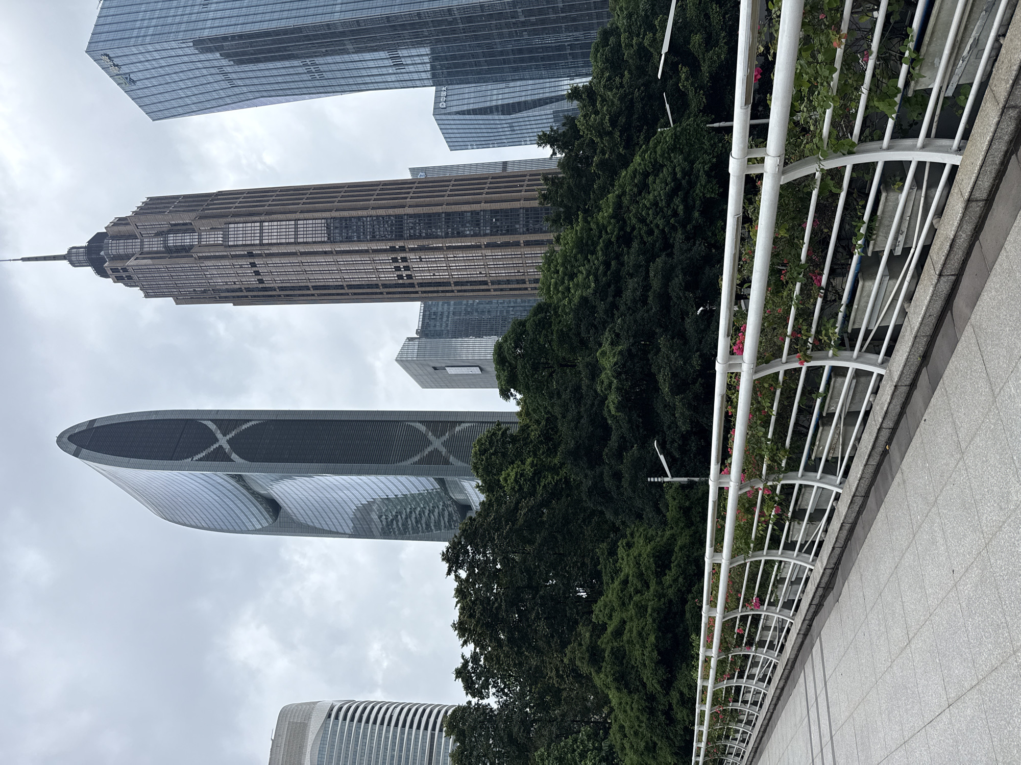 In Tianhe District, China, a pedestrian walkway with a white railing adorned with potted pink flowers overlooks a lush green treeline. Beyond the trees, a cluster of modern skyscrapers pierces the overcast sky. Among the prominent buildings is a tall, slender tower with a pointed spire, a curved, dark glass skyscraper with an X-shaped pattern, and several other rectangular glass structures of varying heights and designs. The overall scene depicts an urban landscape characterized by its modern architecture and abundant greenery.