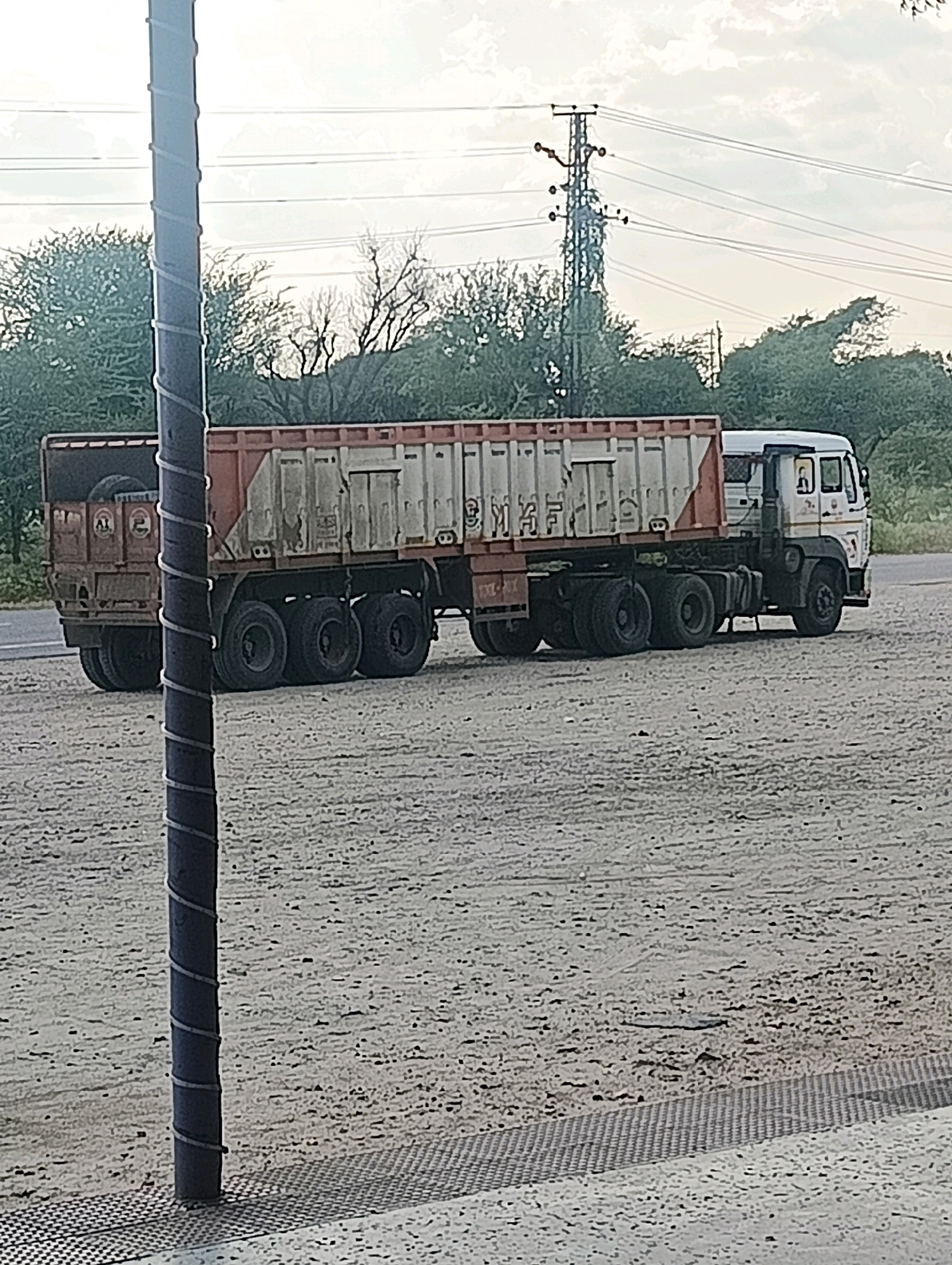 In Chhapri Bari, India, a parked cargo truck sits on a dirt road under an overcast sky. The truck, a long haul vehicle with a white cab and an orange and white cargo container, is positioned to the right of a dark, spiral-wrapped pole in the foreground. The truck's container is weathered, showing signs of use and dirt. Multiple tires are visible, supporting the heavy load. In the background, power lines stretch across the sky, supported by a metal tower. Trees and foliage line the horizon, indicating a rural or semi-rural setting. The ground is covered in gravel and dirt, with a patterned metal grate visible in the lower right corner of the image.