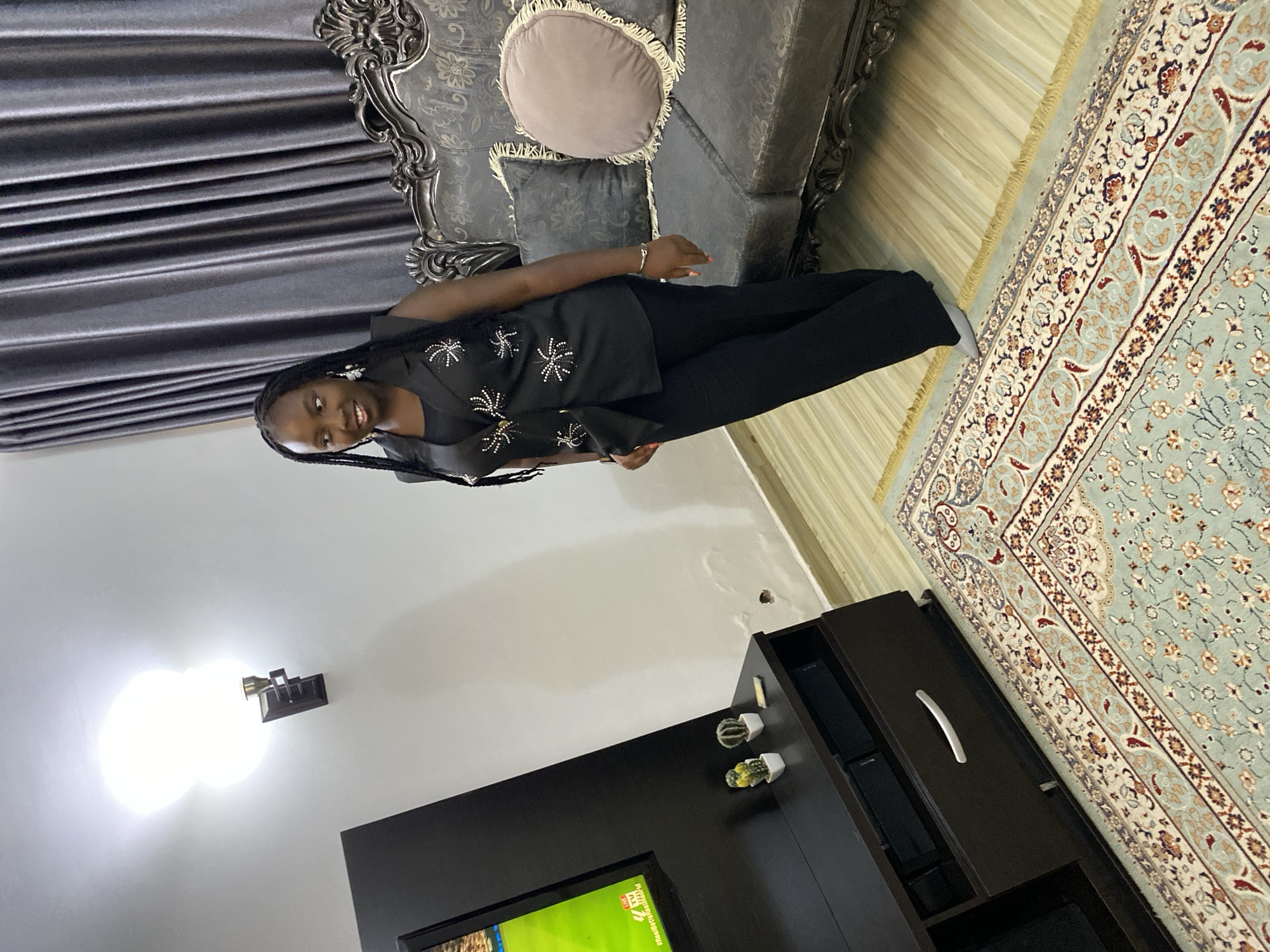 A young woman with dark skin and braided hair is posing indoors in what appears to be a living room in Jos, Nigeria. She is smiling faintly and looking directly at the camera. She wears a black sleeveless top adorned with white and silver star-like embellishments, paired with black trousers. A subtle silver bracelet is visible on her left wrist.
Behind her, to the left, are dark gray pleated curtains. To her right and behind, there's a large, ornate sofa with a dark, patterned fabric and a carved wooden frame. A round, light-colored cushion with fringe and a darker square cushion rest on the sofa.
The floor features a decorative rug in shades of light blue, cream, and reddish-brown, exhibiting intricate floral and geometric patterns. Part of a lighter, possibly wooden or tiled, floor is also visible next to the rug.
To the right of the frame, a dark brown or black TV stand or cabinet is present. On its top surface, two small potted artificial succulent plants serve as decoration. A bright light fixture is mounted on the wall above the cabinet, casting a prominent glow. No discernible text is visible in the image.