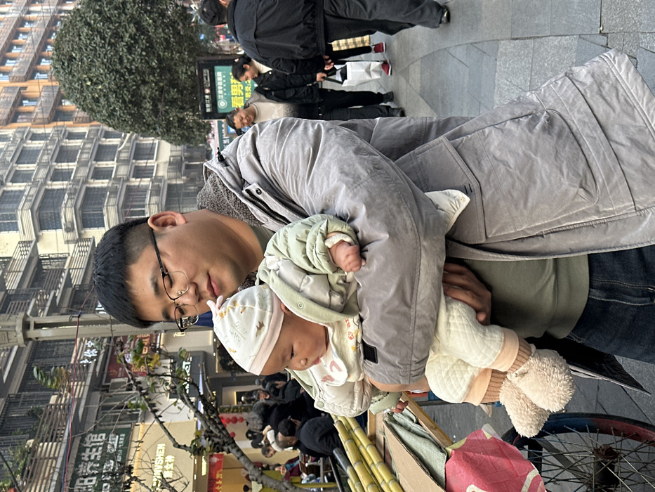 An East Asian adult male is depicted holding an East Asian infant on a paved city street in Jiangjin District, China. The male, appearing in his late 20s to early 30s, has short dark hair, wears dark-framed glasses, a light olive green shirt, a grey padded jacket with a fleecy collar, and dark blue jeans. He is looking down at the infant, who is dressed in a light-colored beanie, a light green padded jacket, a white patterned bib, cream-colored quilted overalls, and light brown fluffy booties. The infant's gaze is directed slightly to its left. To the left of the male, a portion of a vendor's cart with a visible bicycle wheel holds several stalks of sugarcane covered by cardboard. The background features multi-story urban buildings with balconies and visible Chinese signage, including "蒋申医生馆" (Jiang Shen Doctor's Clinic). On the right, a green public sign explicitly displays "江津区" (Jiangjin District) and "江津人才" (Jiangjin Talents). Several pedestrians are visible walking on the sidewalk in the midground and background. A large, pruned tree with dense foliage is situated on the right side of the street. The scene is illuminated by natural daylight.