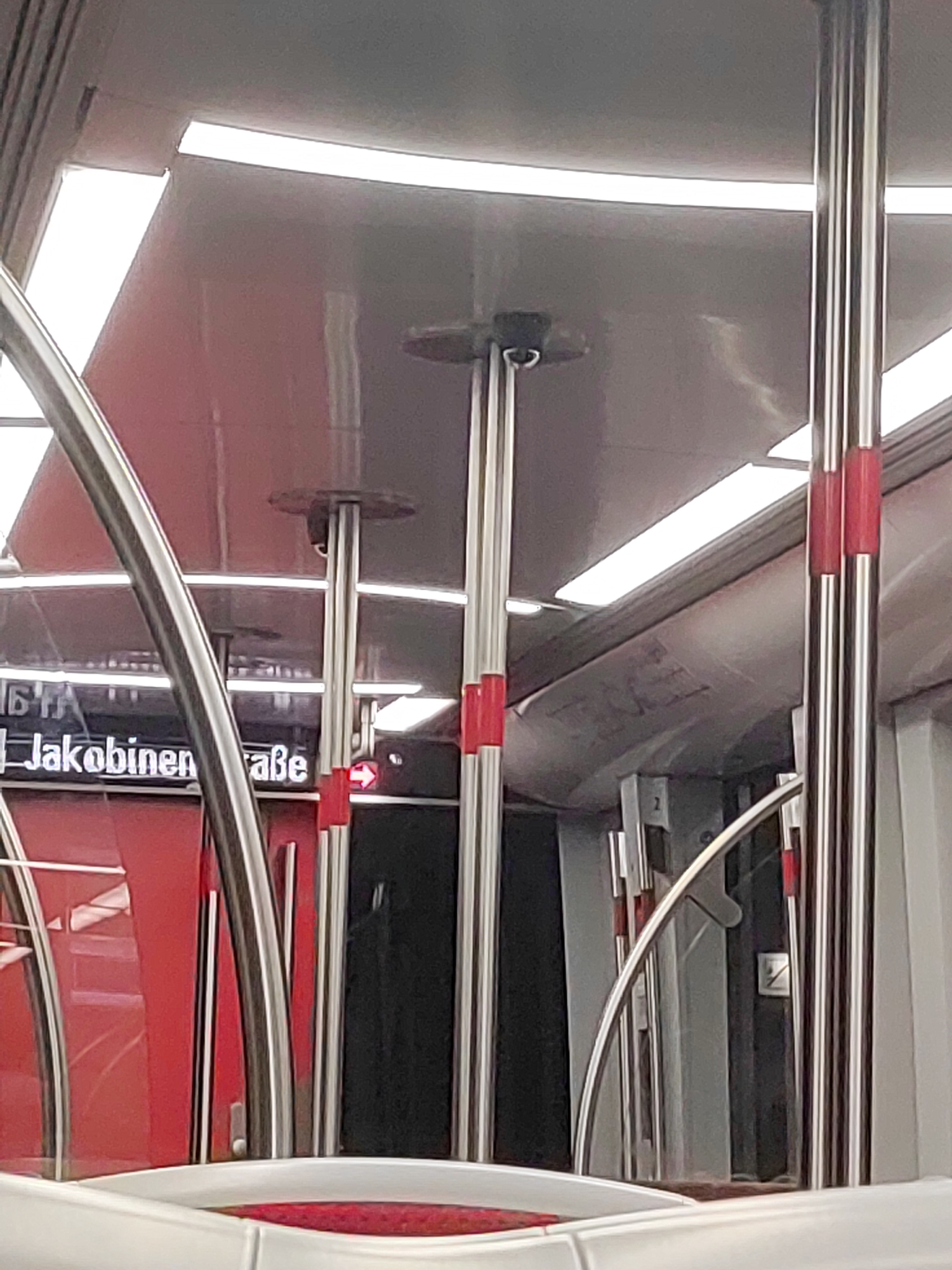 The image depicts the interior of a modern subway train in Fürth, Germany. The scene is characterized by metallic poles, red accents on the walls and poles, and overhead lighting. In the background, a digital display shows the station name "Jakobinenstraße" along with an arrow, indicating it is the current stop. The train car appears to be empty. The overall environment suggests a clean and functional public transportation system.