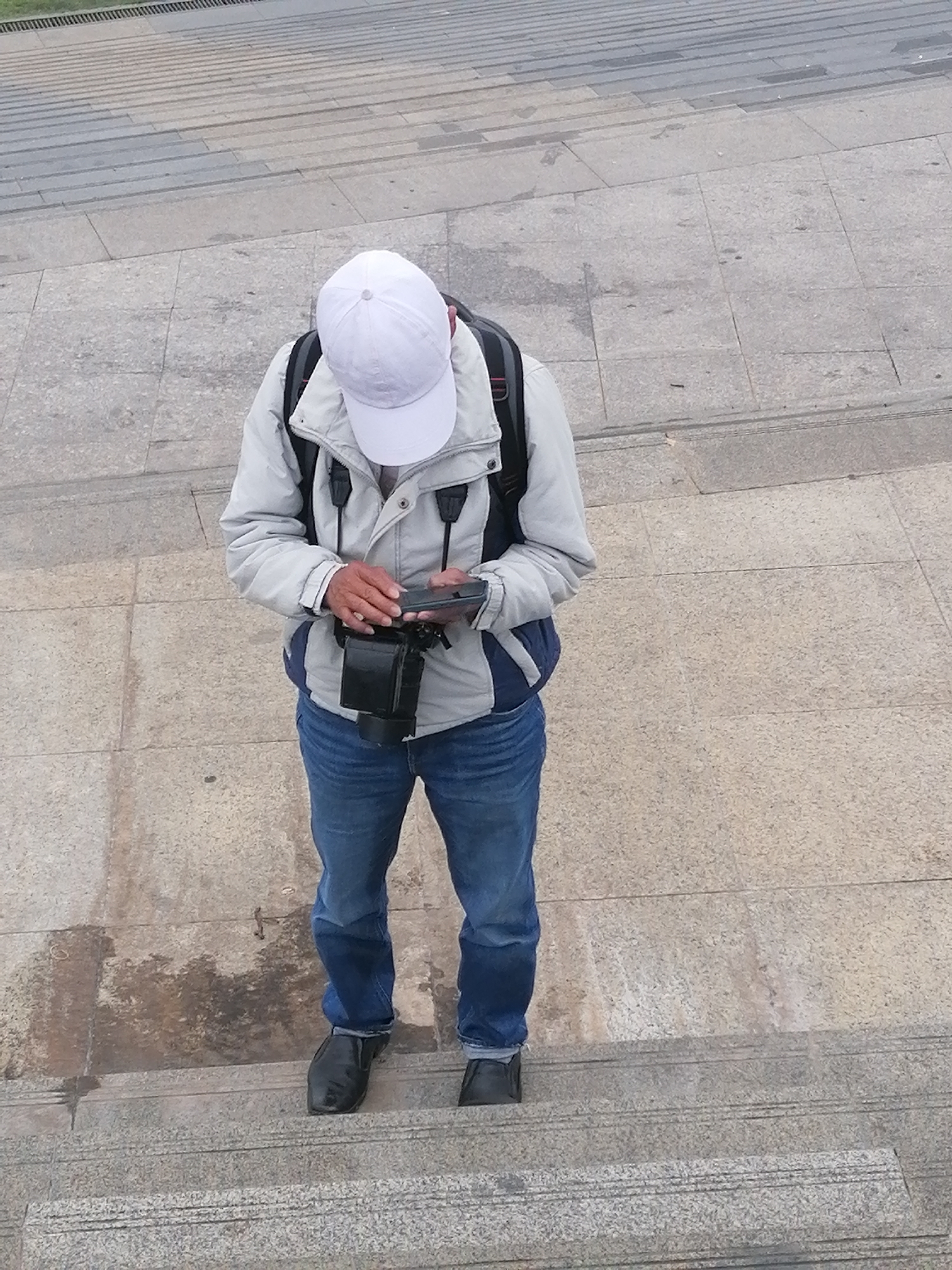 A man wearing a white baseball cap, light grey jacket, blue jeans, and black shoes stands on a grey stone staircase, looking at a mobile phone held in his hands. A camera is slung around his neck, and a backpack is visible on his back. The location is possibly in Da Lat, Vietnam, given the architecture of the staircase and general environment. The man appears to be focused on the phone, possibly checking messages or looking at photos. The background consists of more steps and grey pavement.