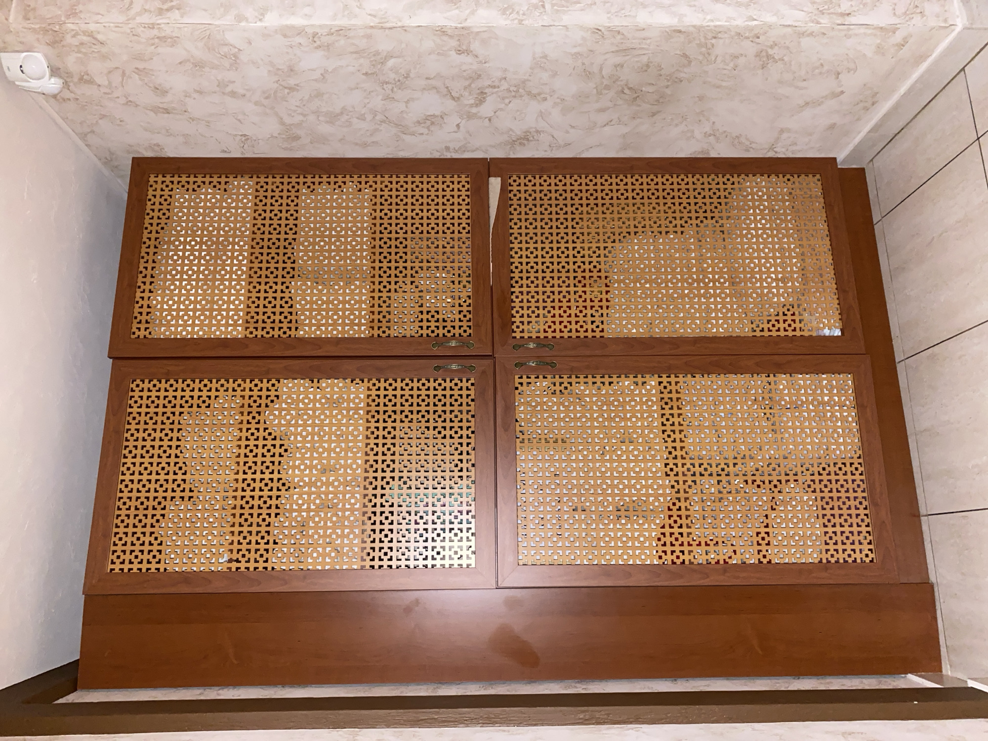 A tall, narrow, four-door wooden cabinet with cane-patterned inserts is shown in a corner of a room. The cabinet has a dark wood finish, and the cane inserts are a light, golden-brown color, with a geometric pattern of small squares. The cabinet doors are divided into two sections, with two doors on the top half and two doors on the bottom half. The cabinet is positioned against a wall with a textured, marble-like wallpaper in shades of cream and brown. A white electrical outlet with a motion sensor is mounted on the wall near the top right corner of the cabinet. The floor is tiled with light-colored square tiles.