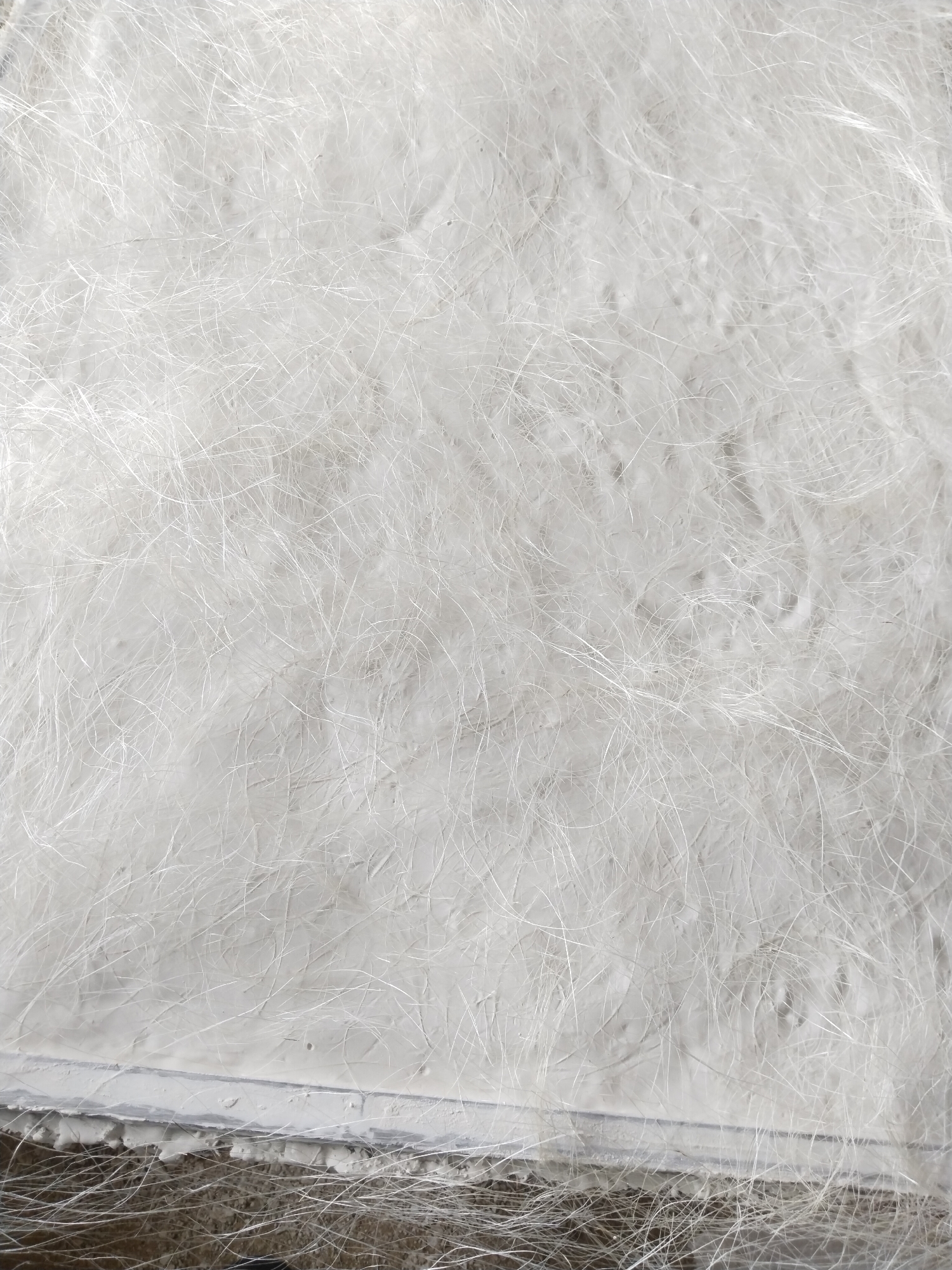 The image shows a close-up, overhead view of a fiberglass mat. The mat is white and composed of a dense network of fine, translucent fibers randomly oriented. The fibers are entangled and appear to be suspended in a matrix that is slightly uneven and textured. The lower portion of the image reveals the edge of the mat, which is supported by a grey, rough surface that may be concrete or asphalt. Some loose fibers extend beyond the edge of the mat. The lighting suggests an outdoor environment, possibly during the day. The scene does not contain any people or recognizable landmarks that would indicate its specific location within Uyo, Nigeria.