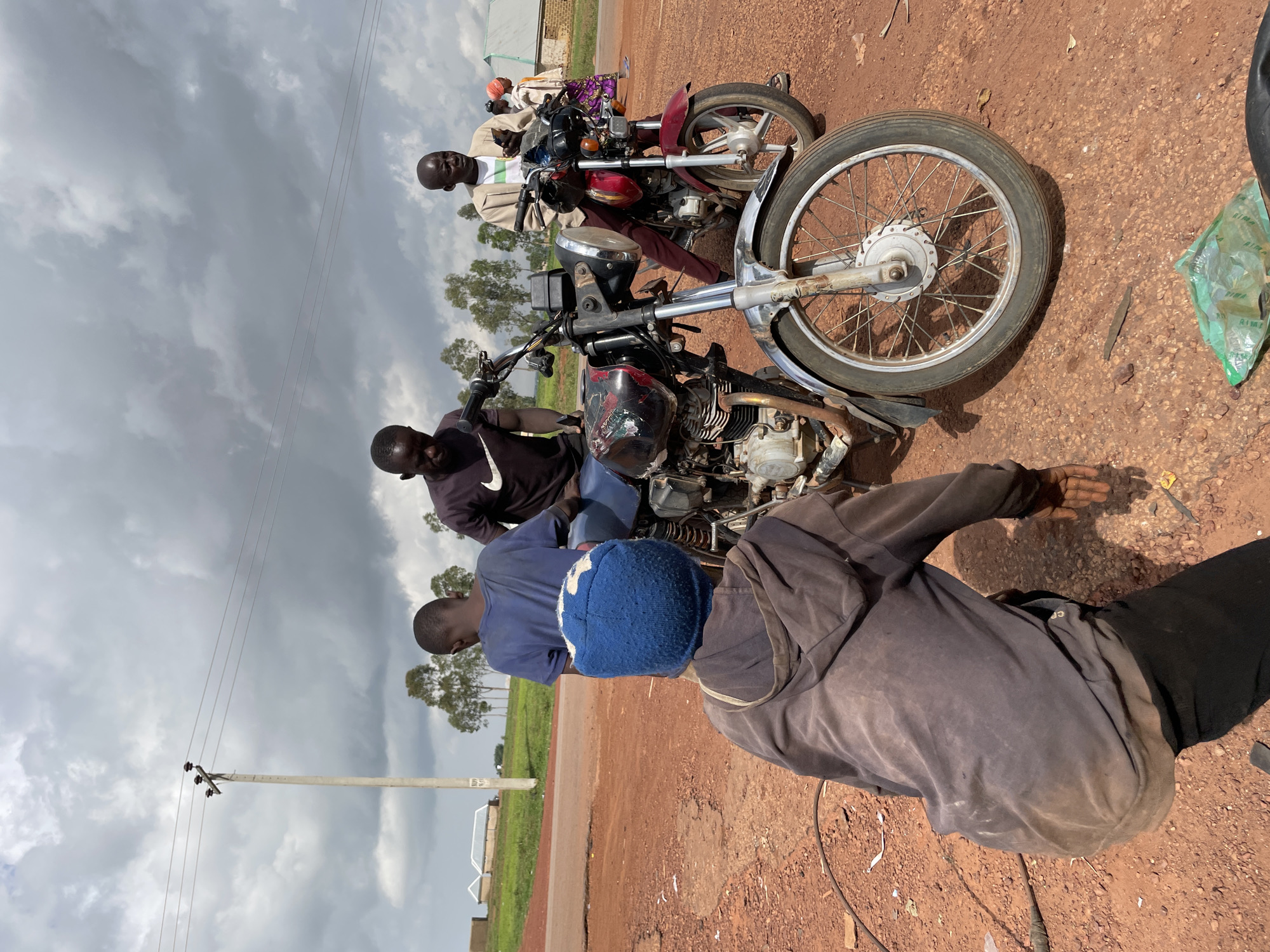 In Wumat, Nigeria, under a cloudy sky, four individuals are gathered around two motorcycles on a dirt road. A young person in a blue knit cap and a grey hoodie is crouched down, facing away from the camera, with their hands near the front wheel of a black motorcycle. To their right, a man in a dark t-shirt stands by the black motorcycle. To the right of the black motorcycle, a second motorcycle, red in color, is parked. A man wearing a light brown jacket over a white shirt stands next to the red motorcycle, looking at his phone. Further to the right, a woman in a colorful patterned dress and a headscarf is visible, also looking towards the motorcycles. In the background, a utility pole with wires stands on a grassy embankment, with some buildings and trees visible further in the distance. The ground is covered in reddish-brown dirt and scattered debris.