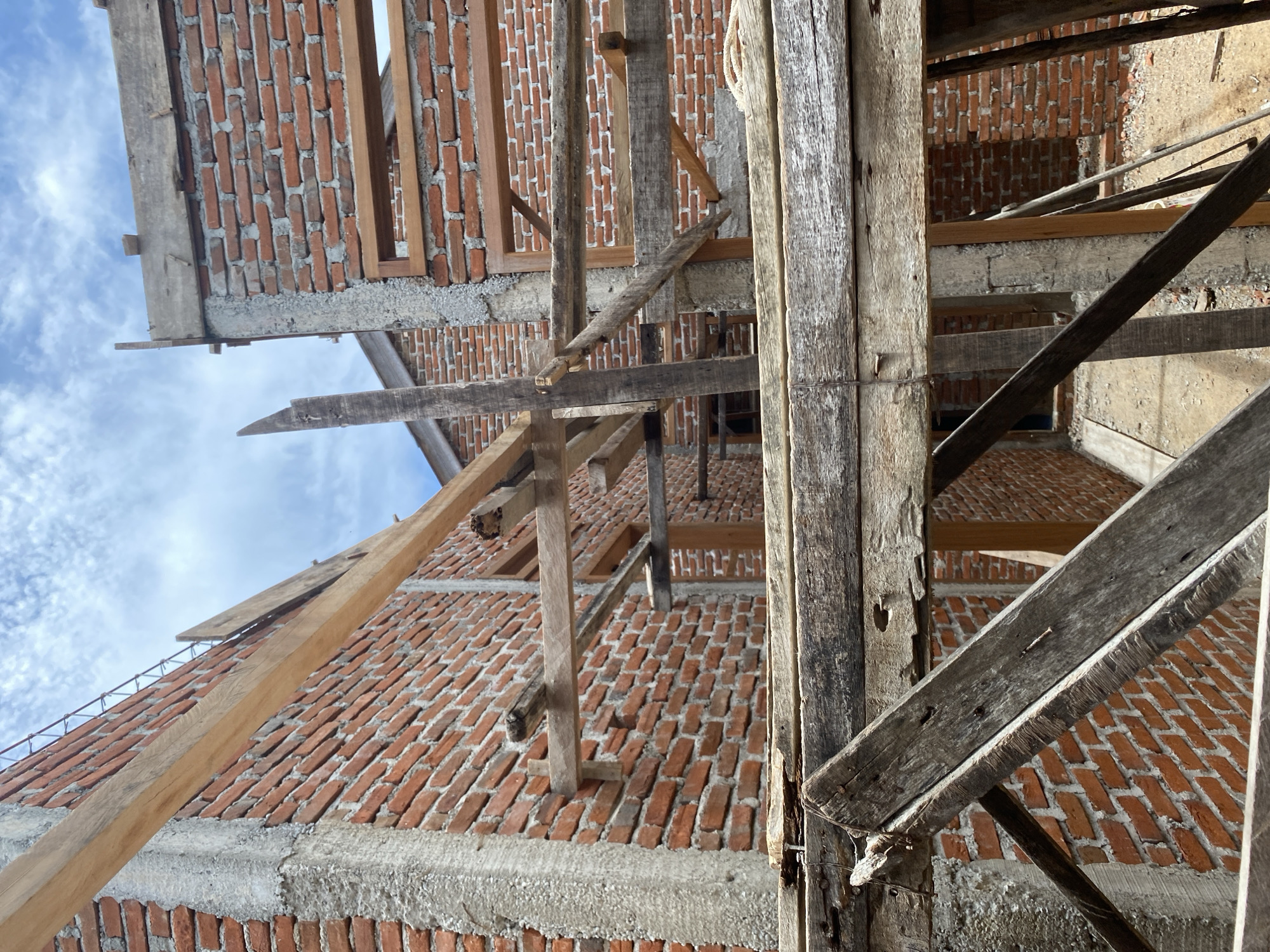 The image captures a close-up, angled view of a building under construction, likely a residential structure. The primary materials visible are unpainted red bricks forming the walls and rough, weathered wooden planks used for scaffolding. Concrete pillars provide structural support. The sky, visible in the upper portion of the frame, is partly cloudy. The scene appears to be outdoors, with debris and loose ground visible in the lower part of the image, suggesting an active construction site. There are no people or animals visible in the frame. The focus is on the architectural elements and the raw materials of construction.