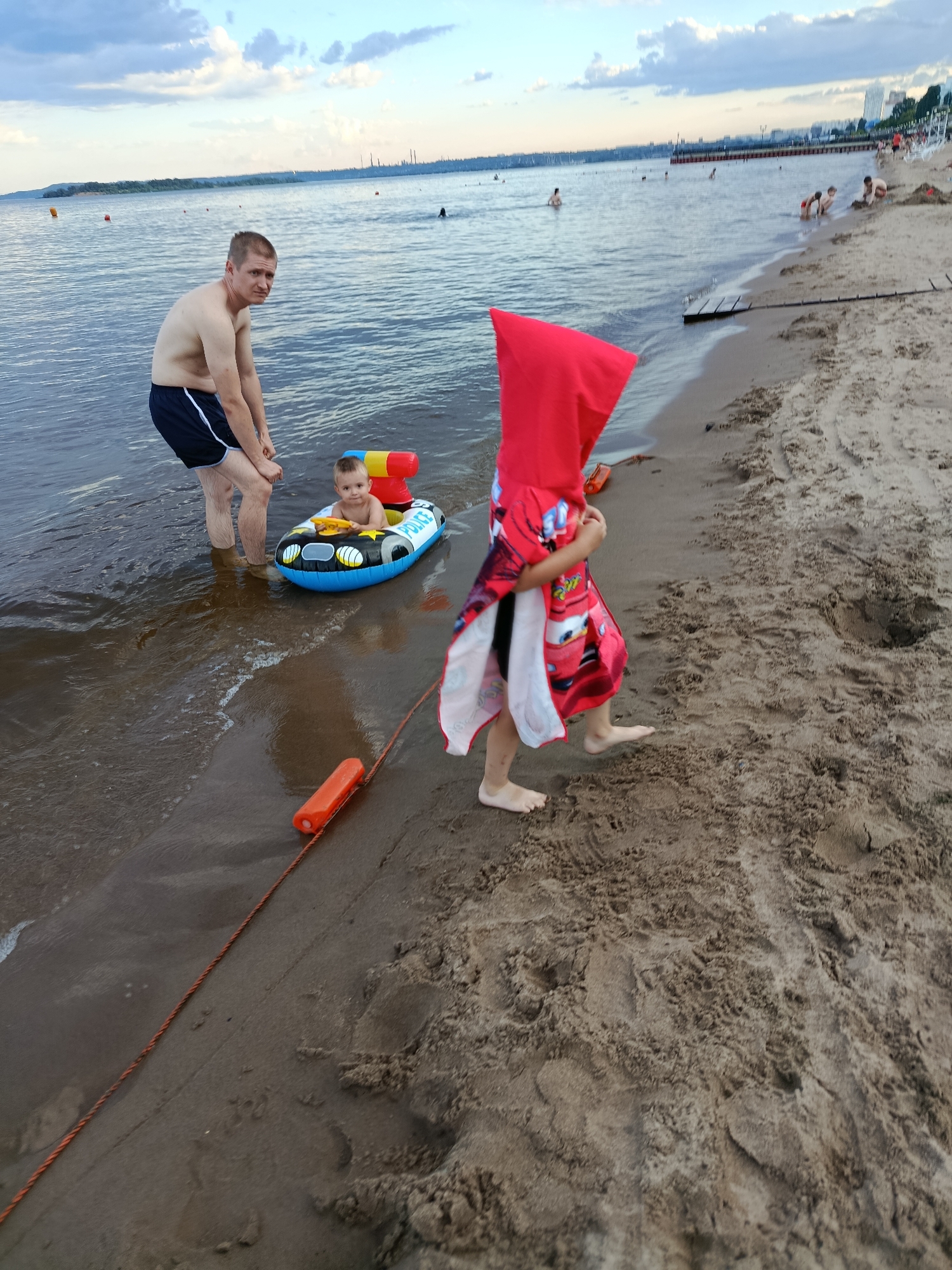 A wide shot captures a sandy beach scene with the Volga River in Saratov, Russia. In the foreground, a man stands knee-deep in the water, looking towards the camera. Next to him, a young child is seated in an inflatable police boat. To the right, another child, partially obscured, is walking on the sand, wearing a red towel or poncho adorned with cartoon characters. An orange float is visible on the sand near the water's edge, connected by a rope. In the background, the river extends towards a distant shoreline with buildings and industrial structures under a cloudy sky. Other beachgoers are scattered along the shore and in the water.