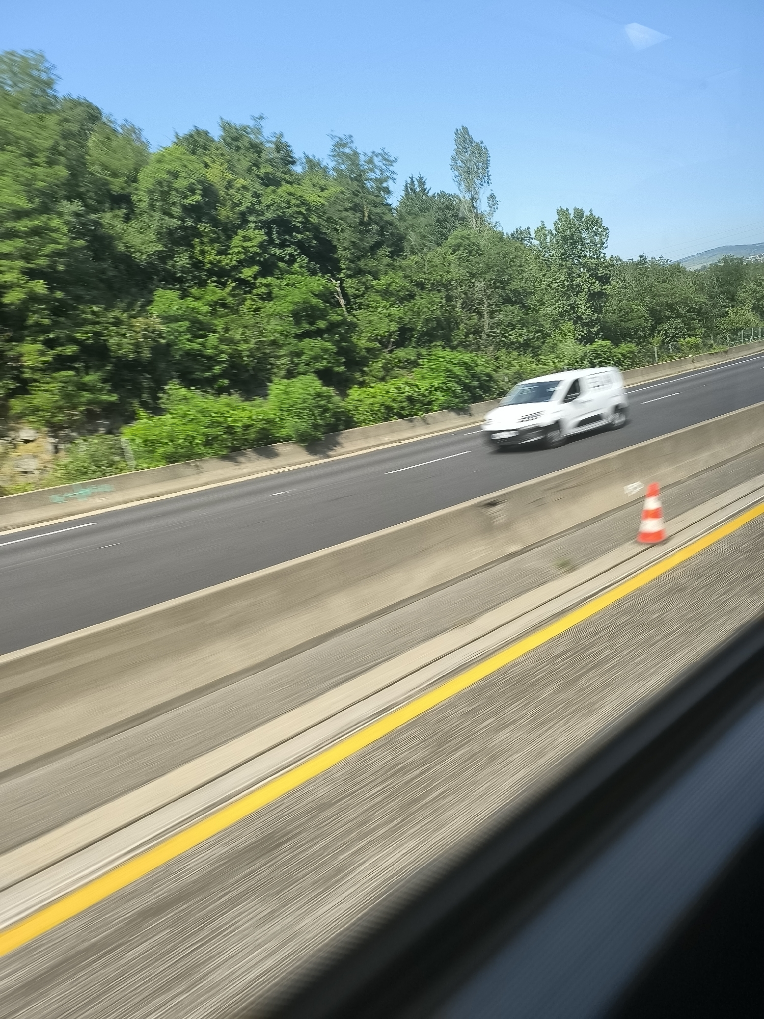 A photograph captured from a moving vehicle depicts a highway scene near L'Horme, France. The road has a concrete barrier on either side, and a white van is traveling in the same direction. A striped traffic cone stands next to the barrier on the right side of the road. The sky is blue and clear, and lush green trees border the highway.