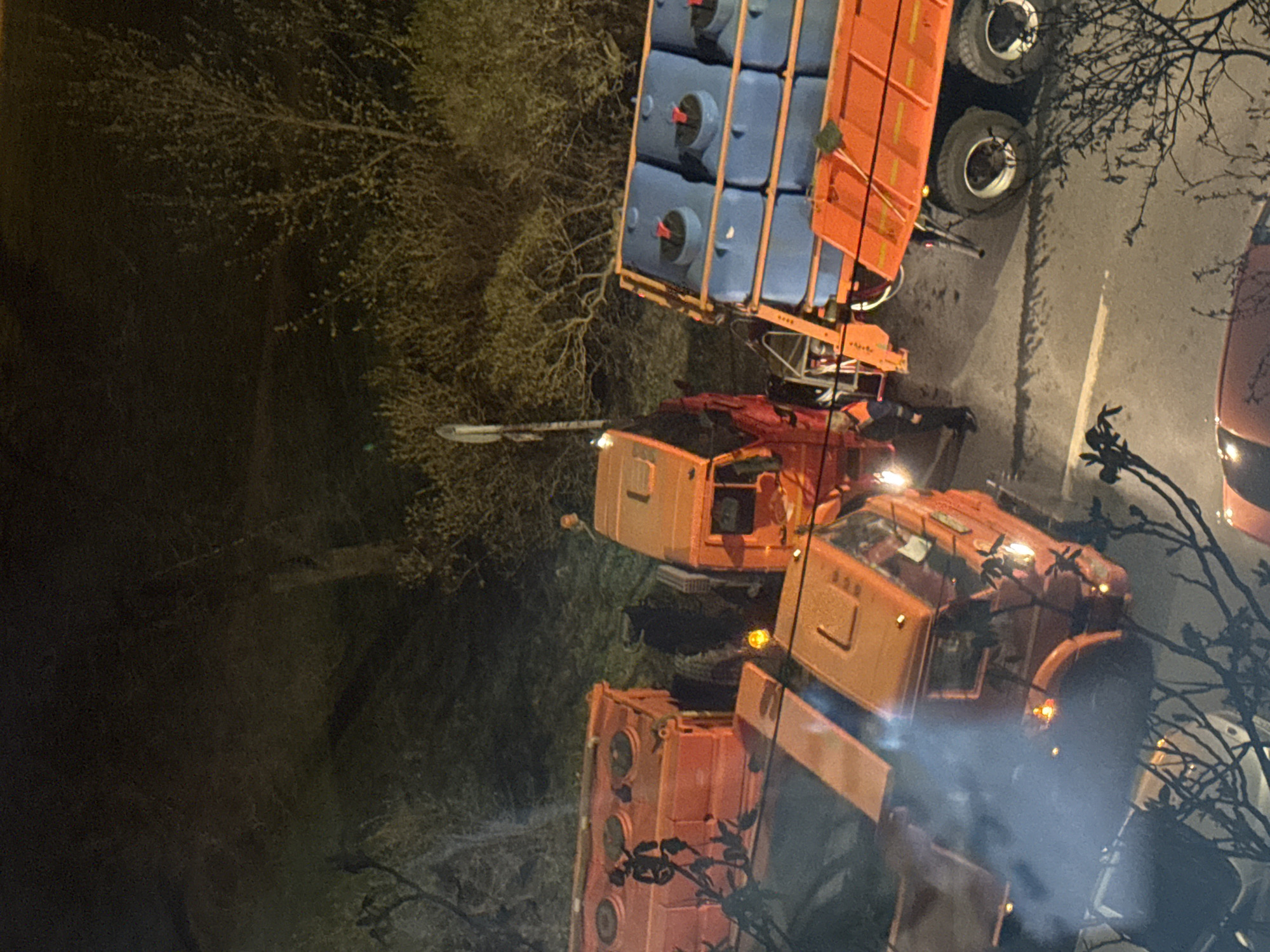 The image, originally oriented vertically, depicts a nighttime scene in what appears to be an industrial or transport area, located in Saint Petersburg, Russia. Several large, orange heavy-duty vehicles are parked on a dark, paved surface, possibly a road or a yard.
In the upper-left section of the image (after rotation), an orange truck is visible, hauling a trailer loaded with numerous blue intermediate bulk containers (IBCs), which are commonly used for transporting liquids or bulk materials. Behind this vehicle, another substantial orange truck, possibly a crane truck or a specialized service vehicle, is positioned. A single person wearing an orange high-visibility vest can be seen standing near this second vehicle, suggesting ongoing work or supervision. A third orange truck is partially visible on the right side of the frame.
The overall setting is dark, illuminated primarily by the vehicles' lights and a distant street light visible on the left. Bare tree branches and sparse vegetation frame the scene, hinting at a late autumn, winter, or early spring season. A light haze or smoke is present near the front of the rightmost truck, possibly exhaust fumes in cold air. No discernible text or explicit location cues other than the provided context are visible in the image. The scene suggests an operation involving heavy transport and logistics during nighttime hours.