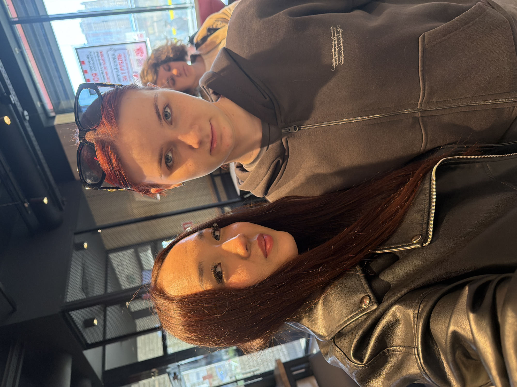 The media file shows a selfie taken by two young women. The woman in the foreground is wearing a black leather jacket and has long, dark brown hair. She is looking towards the camera with a slight smile. Her makeup is noticeable, with dark eyeliner and mascara. The other woman is partially visible behind her, wearing a dark grey hooded sweatshirt. She has red hair and is wearing sunglasses on her head. Her eyes are open and she appears to be looking towards the camera as well. A third person is visible in the background, with curly hair, seemingly asleep.
The setting appears to be indoors, possibly a cafe or a restaurant, with a large window in the background. Sunlight is streaming in, suggesting it is daytime. There is a sign visible through the window with Cyrillic text, and a reflection on a dark surface in the foreground, possibly a table. The overall mood seems casual and friendly.
Visible text includes Cyrillic script on a sign in the background, though it is not clearly legible. The sweatshirt of the woman in the middle ground has a subtle embroidered logo.