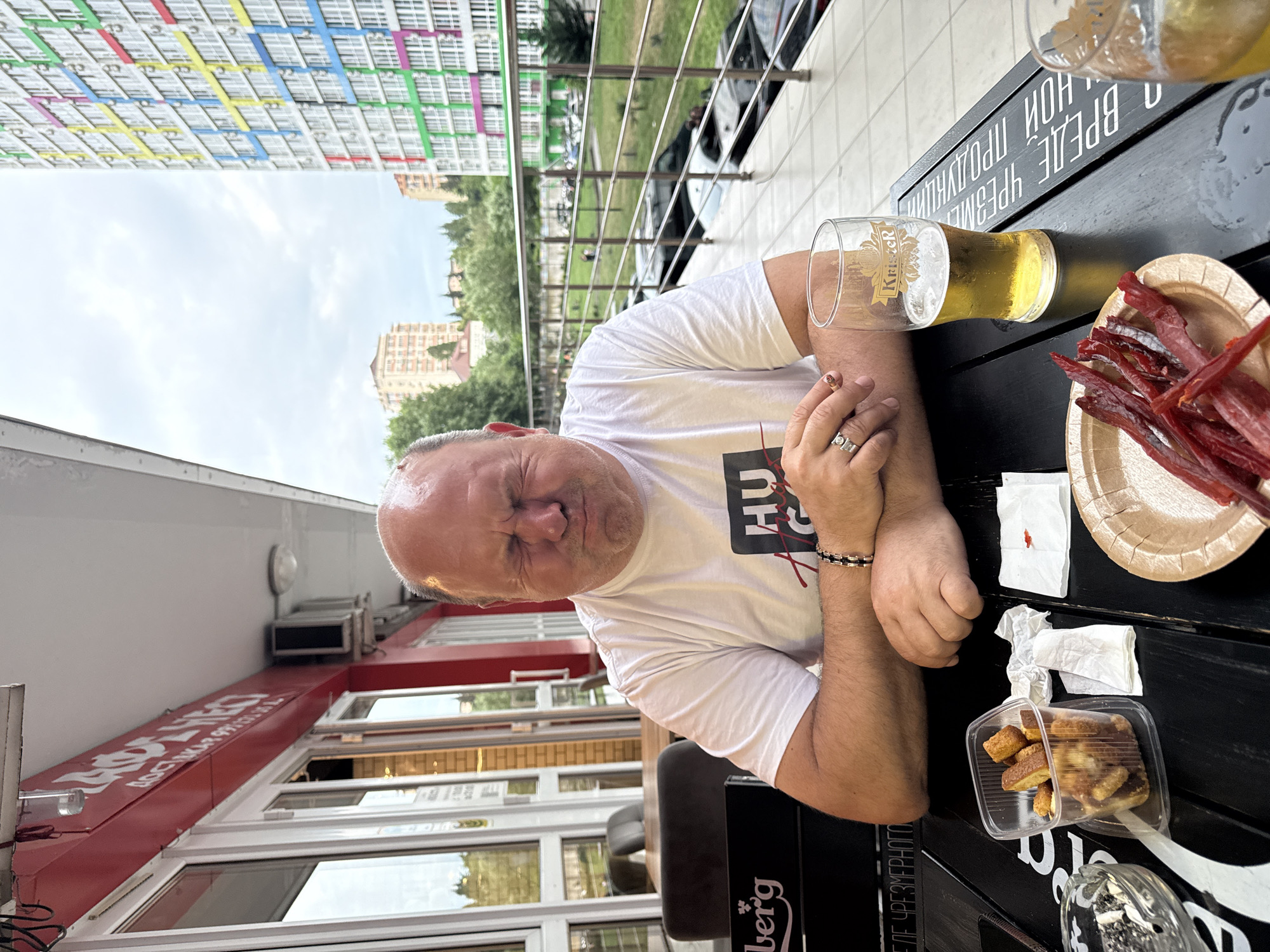 In Sochi, Russia, a middle-aged bald man with his eyes squeezed shut and mouth contorted in a grimace is seated at a black table on an outdoor patio. He wears a white t-shirt with a black graphic and a bracelet on his left wrist, and a ring on his right ring finger. On the table in front of him are a glass of beer, a plate of dried meat sticks, a container of croutons, a crumpled napkin, and an ashtray with cigarette butts. In the background, a modern, colorful apartment building towers over other structures and trees, indicating an urban setting. The railing of the patio is visible, along with parked cars below. The overall ambiance suggests a casual outdoor dining or drinking establishment.