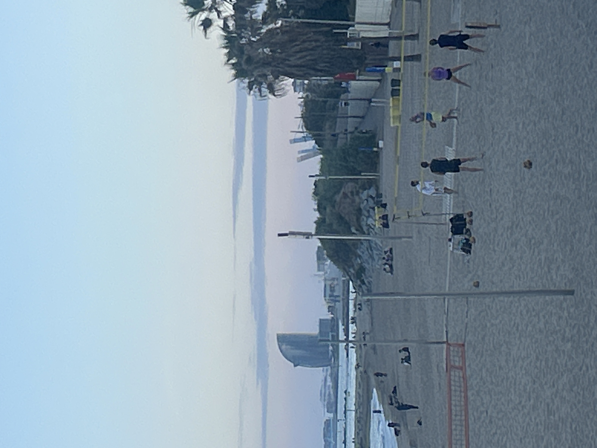 The image depicts Barceloneta Beach in Barcelona, Spain. Several individuals are playing volleyball on the sand near the beach. The Hotel Arts Barcelona is visible in the background near the coast. The sky is light blue with slight cloud cover. Palm trees are scattered along the beach's edge. The general environment is calm, indicative of a late afternoon or early evening setting.
